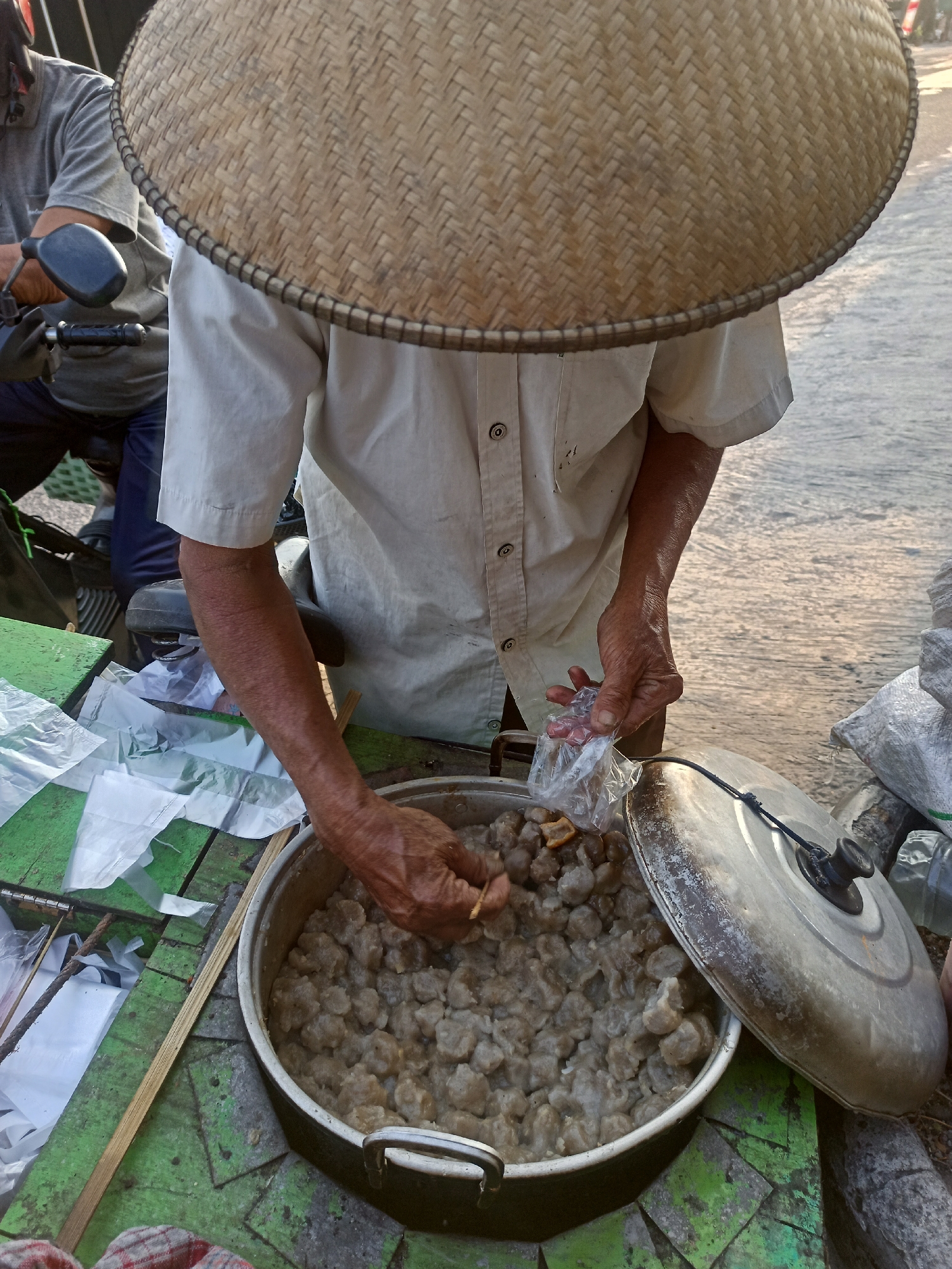 In Gemolong, Indonesia, a street vendor wearing a traditional conical woven hat stands behind a cart. The vendor, dressed in a light-colored collared shirt and light-colored pants, is in the process of serving food. Their right hand is holding a small plastic bag containing food items, while their left hand is reaching into a large pot filled with numerous small, round, light-brown food pieces, possibly meatballs or dumplings. The pot is situated on a green wooden surface, which appears to be the cart. Beside the pot lies a large, weathered metal lid. To the left of the pot, there are several white plastic bags and a portion of another person sitting on a motorcycle, with their hand on the handlebar. The background suggests an outdoor, possibly street setting with a paved surface visible.