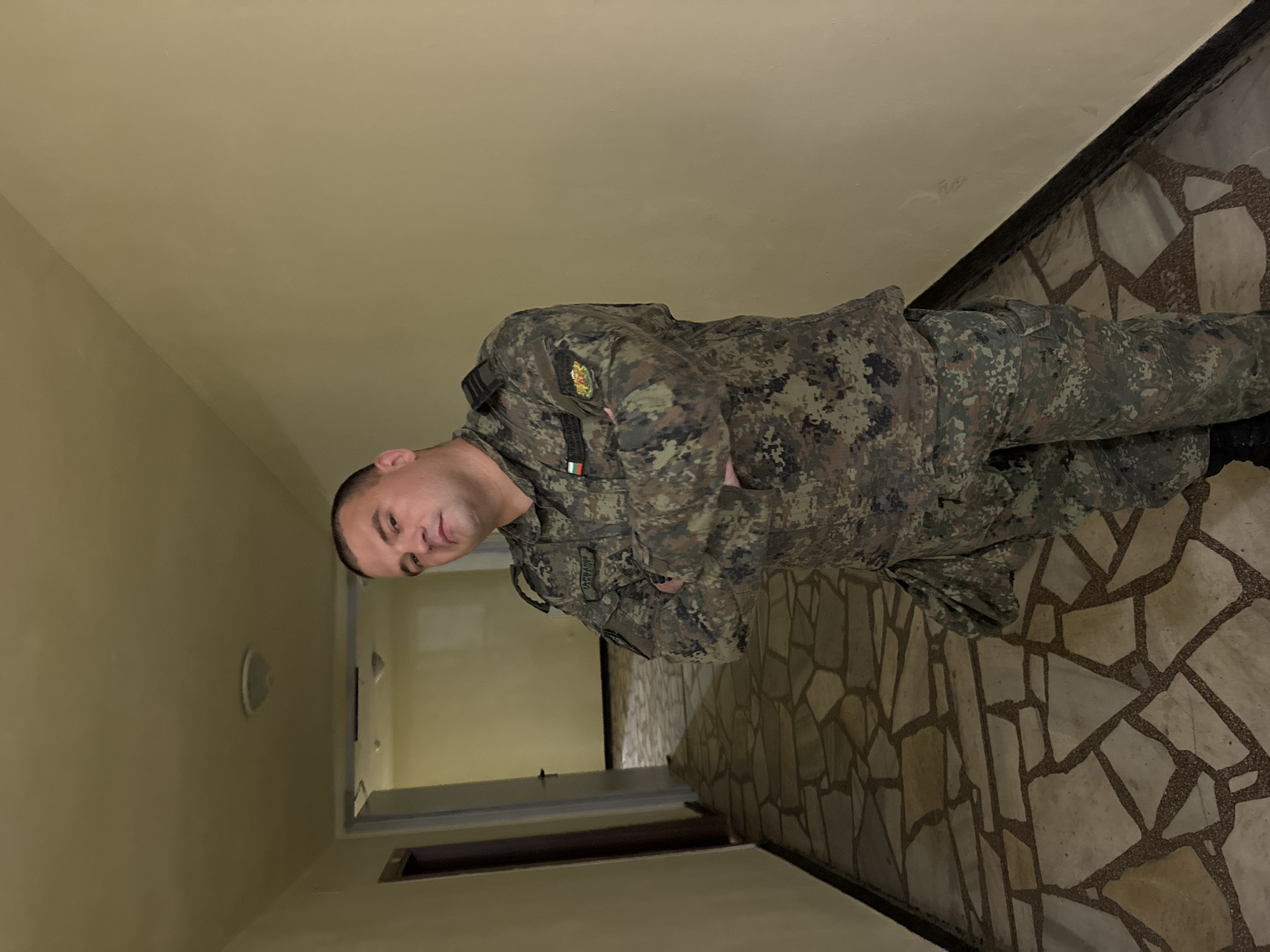 A male individual in a military-style camouflage uniform stands centered in a beige-walled indoor hallway. The uniform features a digital camouflage pattern with shades of green, brown, and grey. A name tape displaying "ЗАБАРГАНОВ" in Cyrillillic is visible on the wearer's right chest. A rectangular patch depicting the Bulgarian flag is on the right shoulder, and a circular patch resembling the coat of arms of Bulgaria is on the left shoulder. Rank insignia, consisting of two black bars, is present on both epaulets. The individual has short hair and a neutral expression, looking directly at the camera with arms crossed over the chest. The hallway floor is paved with irregularly shaped light-colored stone or tile pieces interspersed with darker grout. The walls are solid, light beige, and the ceiling is also light-colored with visible circular light fixtures. Two doorways are visible in the background further down the corridor. The scene is illuminated by artificial light. The location is within a building in Unknown, Unknown.