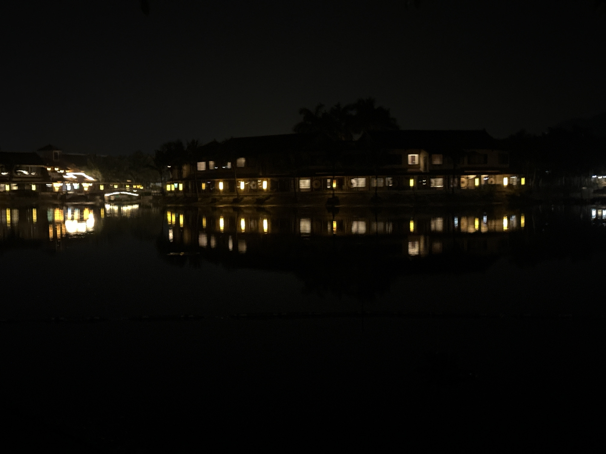In the city of Nam Hoa Lư Ward, Vietnam, a dimly lit scene unfolds by a body of water at night. The image captures a row of buildings with illuminated windows reflecting on the dark water's surface. The architecture appears to be traditional, with sloped roofs and multiple stories. Trees and foliage are visible behind the buildings, silhouetted against the night sky, with some palm trees suggested. A small, arched bridge connects sections of the buildings or land. The reflections on the water are distorted, elongated streaks of light, indicating a slightly rippled surface. The overall ambiance is serene and somewhat mysterious, characteristic of a quiet evening in a waterside location.