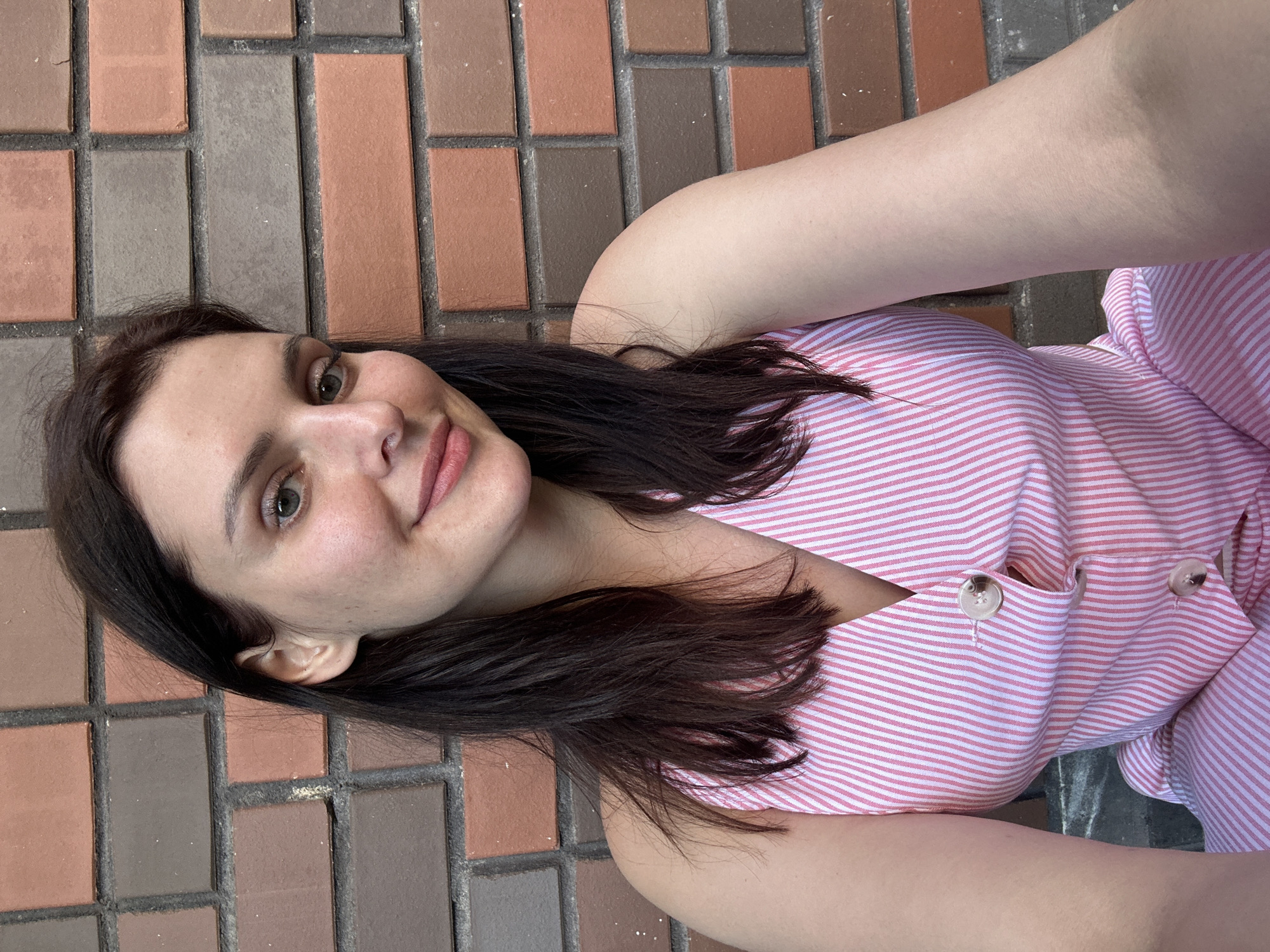 A young woman lies on a brick-paved surface, her head angled towards the camera. She has dark hair that falls around her shoulders and is wearing a pink and white striped shirt. The shirt has two buttons visible near the neckline. The brick paving is made up of rectangular bricks in various shades of brown and grey. The lighting suggests it is daytime, possibly outdoors, with a soft, natural light. The woman's expression is relaxed, with a slight smile. The scene is a close-up shot, focusing on the woman's face and upper body, with the brickwork serving as the background.
