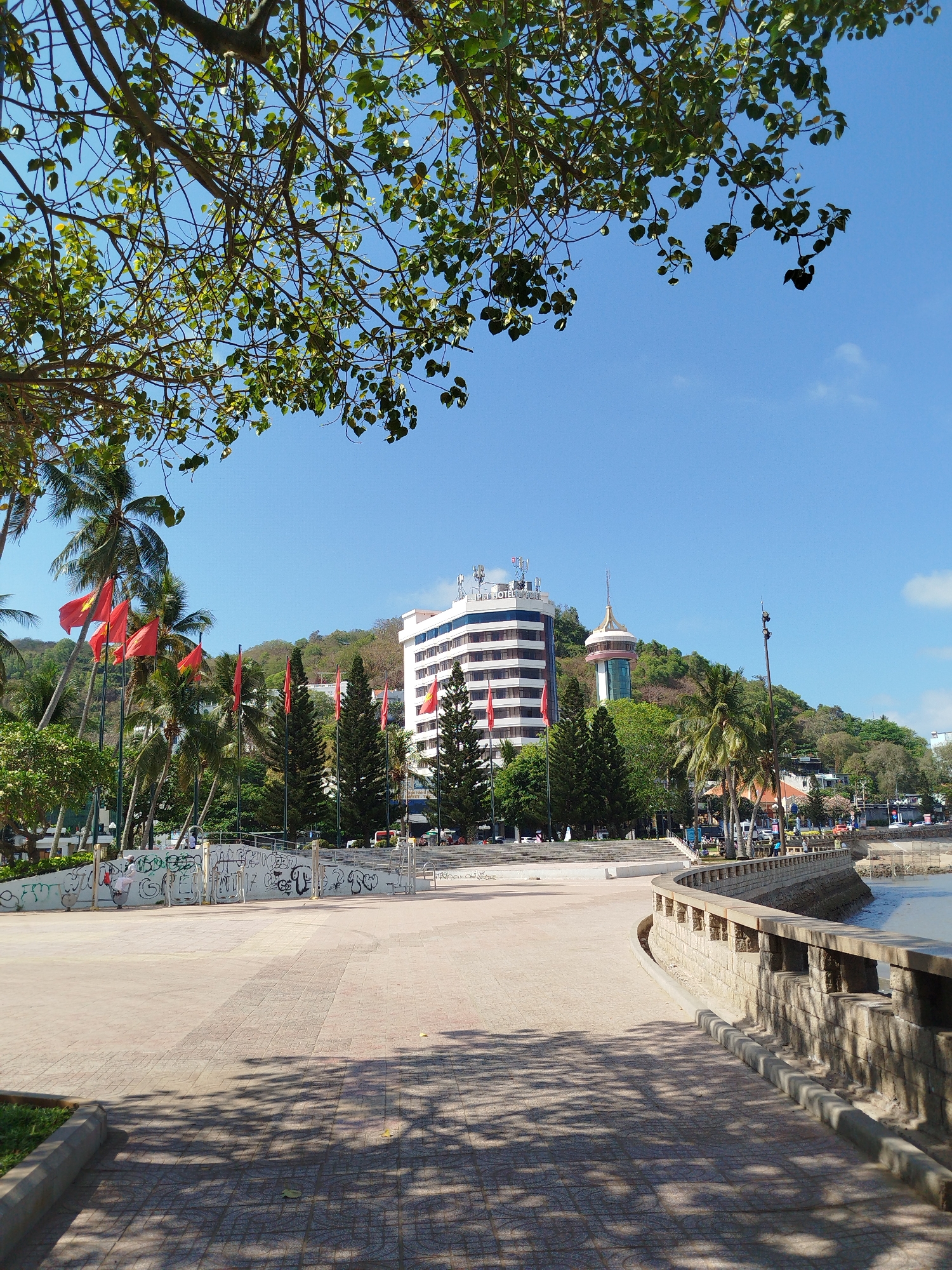 A daytime outdoor shot presents a landscape in what appears to be Vung Tau, Vietnam. In the foreground, there is a tiled pedestrian walkway with shadows from overhead trees creating a dappled effect. A stone railing runs along the right side, bordering a body of water.
In the mid-ground, there is a large open area with a low wall covered in graffiti. Beyond this area, there are palm trees and several Vietnamese flags fluttering in the breeze. Behind the trees, there is a large, white high-rise building and a tower atop a lush, green hill.
The sky above is clear and blue with scattered leaves from an overhanging tree branch along the top of the frame. There are no visible people in the immediate foreground, but there are indistinct figures in the distance.