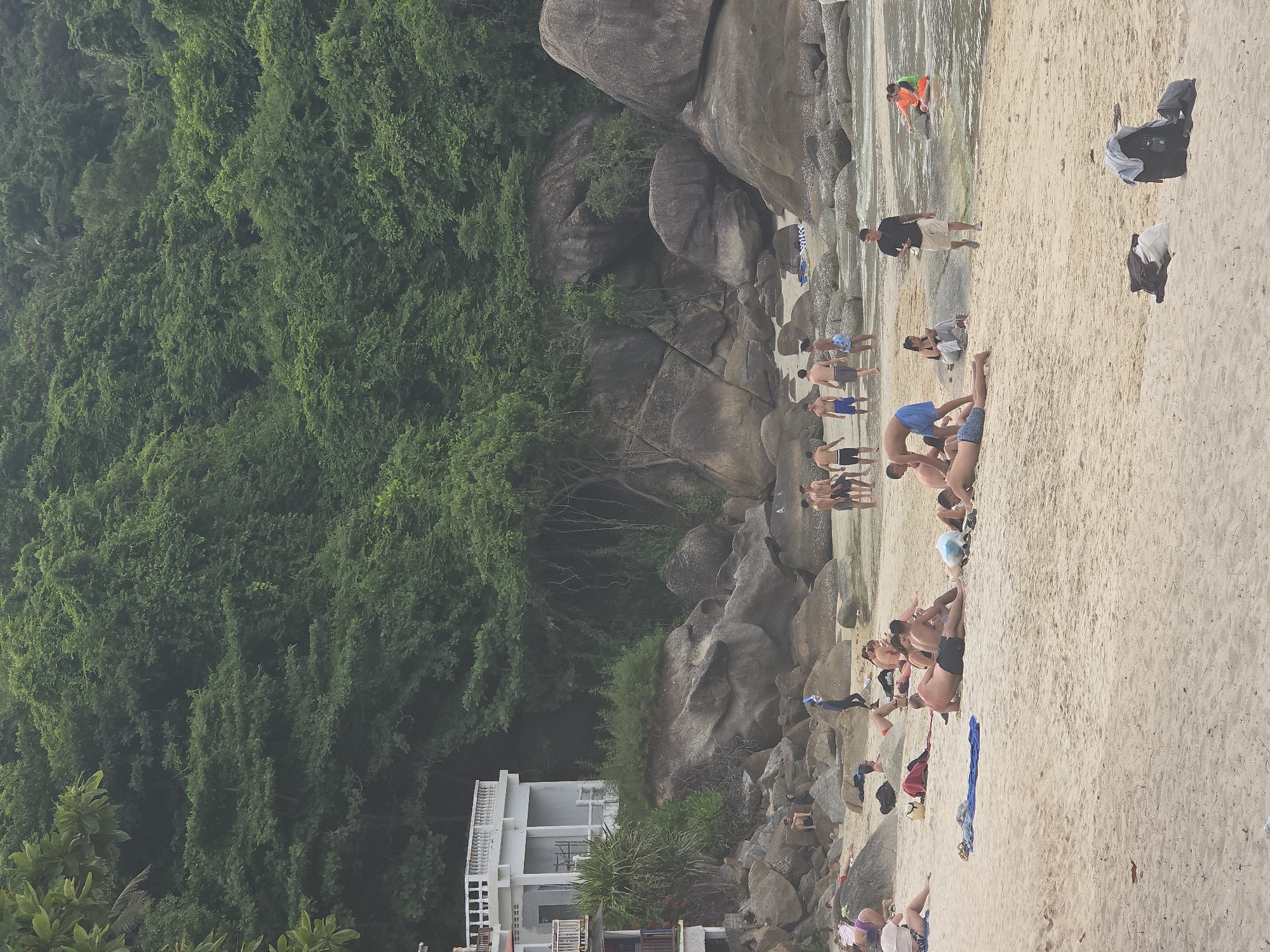 The image depicts a crowded beach scene with numerous people engaged in various activities. The beach is sandy and appears to be located in a tropical or subtropical region, given the lush green vegetation covering a rocky hillside in the background. Several large boulders are scattered on the shore. People are sunbathing, sitting, and standing on the sand. Some individuals are in the shallow water, with one person in an orange and green outfit appearing to be playing or wading. In the mid-ground, a group of men are standing and interacting, while others are lying down on towels. In the foreground, several people are also relaxing on the sand. A white building with balconies and railings is visible on the left side of the frame, partially obscured by trees and vegetation. Bags and towels are scattered on the sand throughout the scene. The sky is overcast, suggesting a diffused light condition.