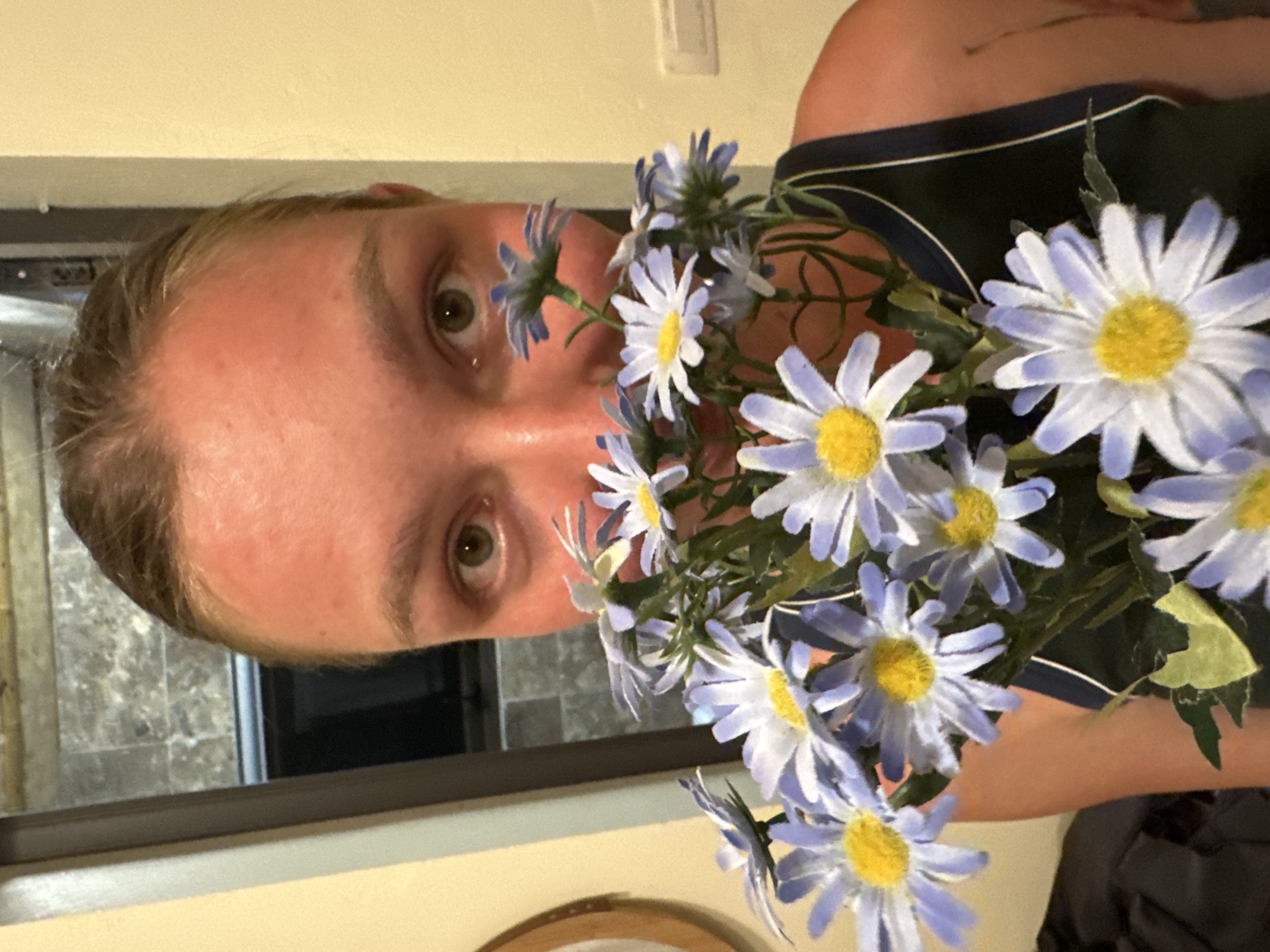 A person with light brown hair, pulled back, holds a bouquet of artificial daisy-like flowers in front of their face, obscuring their mouth and lower nose. The flowers have light blue-purple petals and bright yellow centers, with green stems and artificial green leaves. The person's forehead, nose bridge, and cheeks display a reddish tone, consistent with sunburn. Their green-brown eyes gaze directly forward. They are wearing a dark-colored tank top with a thin white trim along the neckline or strap.
The background consists primarily of light yellow walls. To the left of the person, a partial dark circular wooden frame, possibly from a mirror or decorative item, is visible. Behind the person's head, a dark-framed opening, appearing to be a window or doorway, reveals a darker interior space with a patterned, possibly tiled, dark grey surface. To the right, a white rectangular electrical outlet or switch plate is mounted on the wall. The image is attributed to the city of Ta Xua, Vietnam.