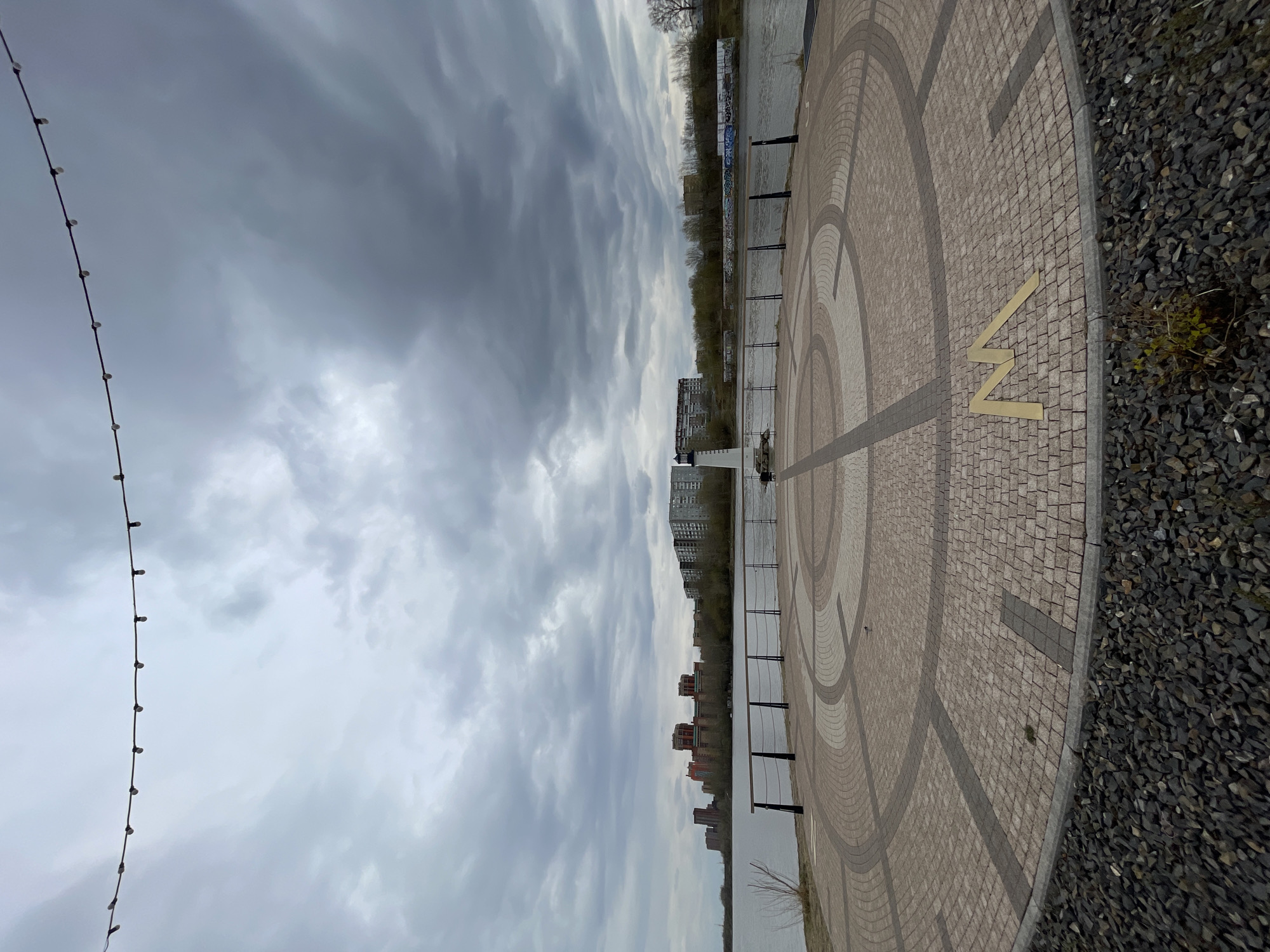 The image shows a section of the embankment along the Yenisey River in Krasnoyarsk, Russia. The foreground features a paved area with a circular pattern and a large "N" symbol inlaid in brass. To the right of the paved area is a section of gravel. A string of lights is visible on the left. In the background, across the river, are several buildings, a strip of trees, and a cloudy sky. The river runs between the embankment and the buildings. A metal railing runs along the edge of the embankment.