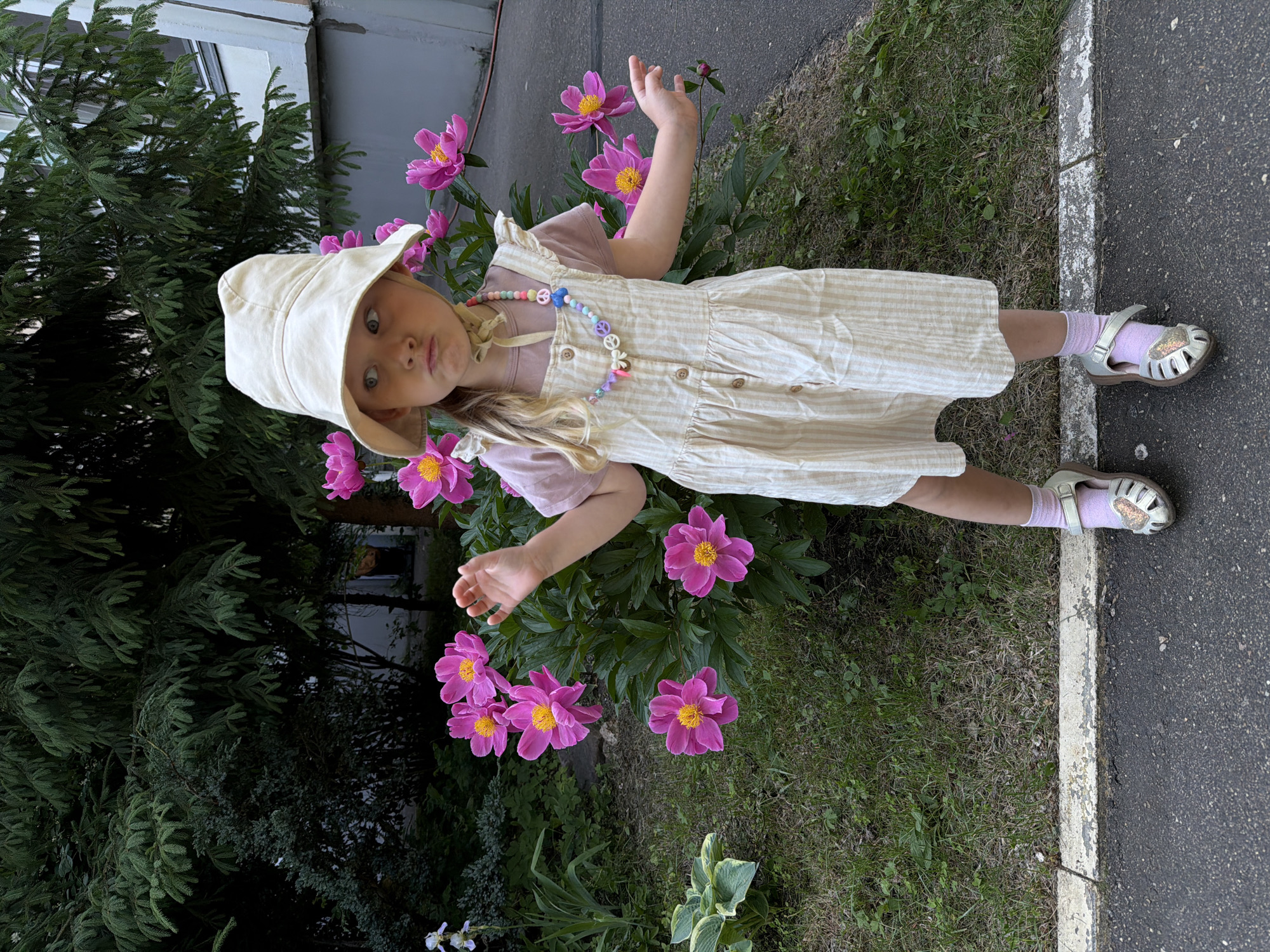 A young girl wearing a beige bucket hat, a pink t-shirt, and a striped beige pinafore dress stands in front of a bush with pink peonies. She is also wearing pink socks and silver sandals. A beaded necklace with peace signs and colorful beads is around her neck. She is looking to her left with her mouth pouted. The background shows green trees, a building, and a paved area. This is likely an urban environment, possibly a courtyard or park in Moscow, Russia.