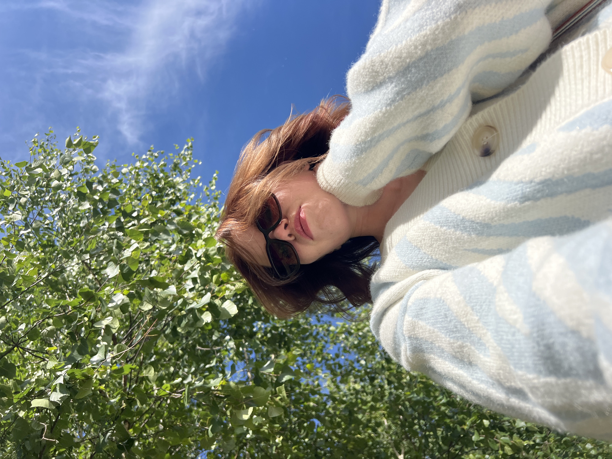 A woman with auburn hair and sunglasses wears a blue and white striped cardigan while standing outdoors, with a clear blue sky and trees with green leaves visible in the background. The perspective is from a low angle, looking up at the woman. There is no information available regarding the location of the image.