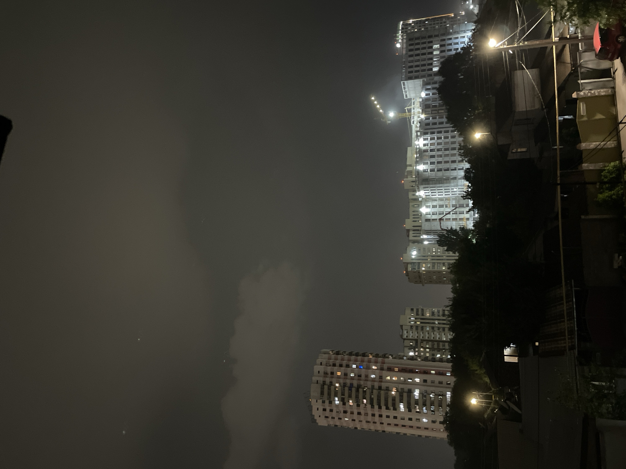 The image depicts a night scene in Almaty, Kazakhstan, featuring a cluster of buildings and a construction site against a dark, cloudy sky. In the foreground, the roofs of low-rise structures and a fence are visible, with a small red car parked on the right. Behind these, a line of trees partially obscures the view of more distant buildings. Dominating the midground and background are several multi-story residential buildings, illuminated with lights from their windows. One tall, circular building stands out on the left, while a long, multi-sectioned building complex, appearing to be under construction, stretches across the center and right of the frame. A construction crane is prominently visible above this development. The overall atmosphere is urban and nocturnal.
