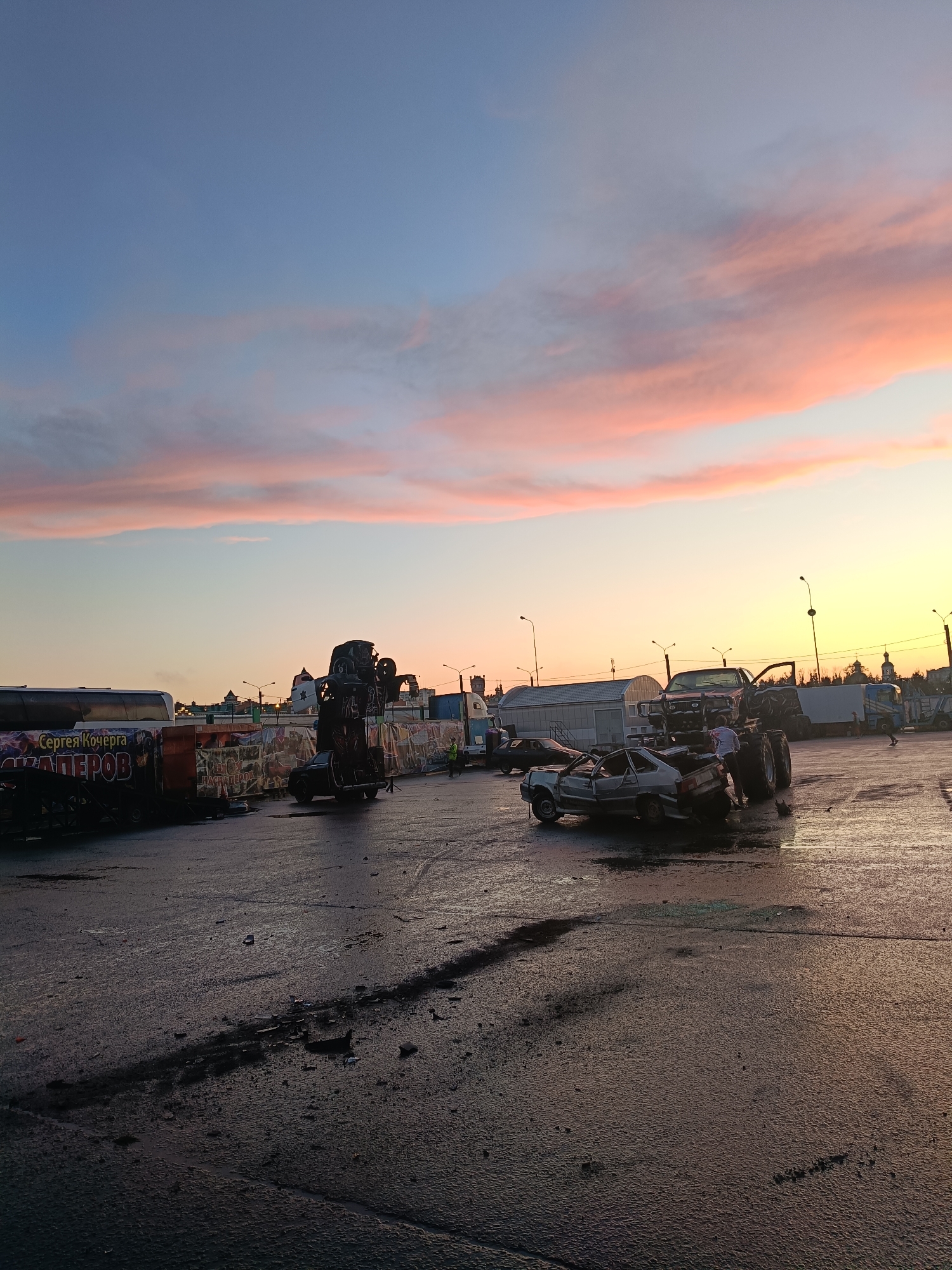 The image depicts an outdoor scene at dusk in the city of Yoshkar-Ola, Russia, possibly a car stunt arena or a location after an event. The sky is filled with colorful clouds in shades of pink, orange, and blue, indicating sunset. The ground appears to be wet asphalt, with tire marks and debris scattered across it.
In the foreground and midground, several wrecked and modified vehicles are present. One notable vehicle is a monster truck with large tires, parked near smaller, crushed cars. Another vehicle is overturned, its wheels in the air, positioned to the left of the monster truck. A large trailer with banners advertising "Сергей Кочерга" and "МАСТЕРОВ" is visible on the left side of the frame. Several people, dressed in casual attire and some in safety vests, are observing the scene or interacting with the vehicles. In the background, buildings and streetlights suggest an urban environment. The overall impression is one of a post-event atmosphere, likely related to a car show, stunt performance, or demolition derby.