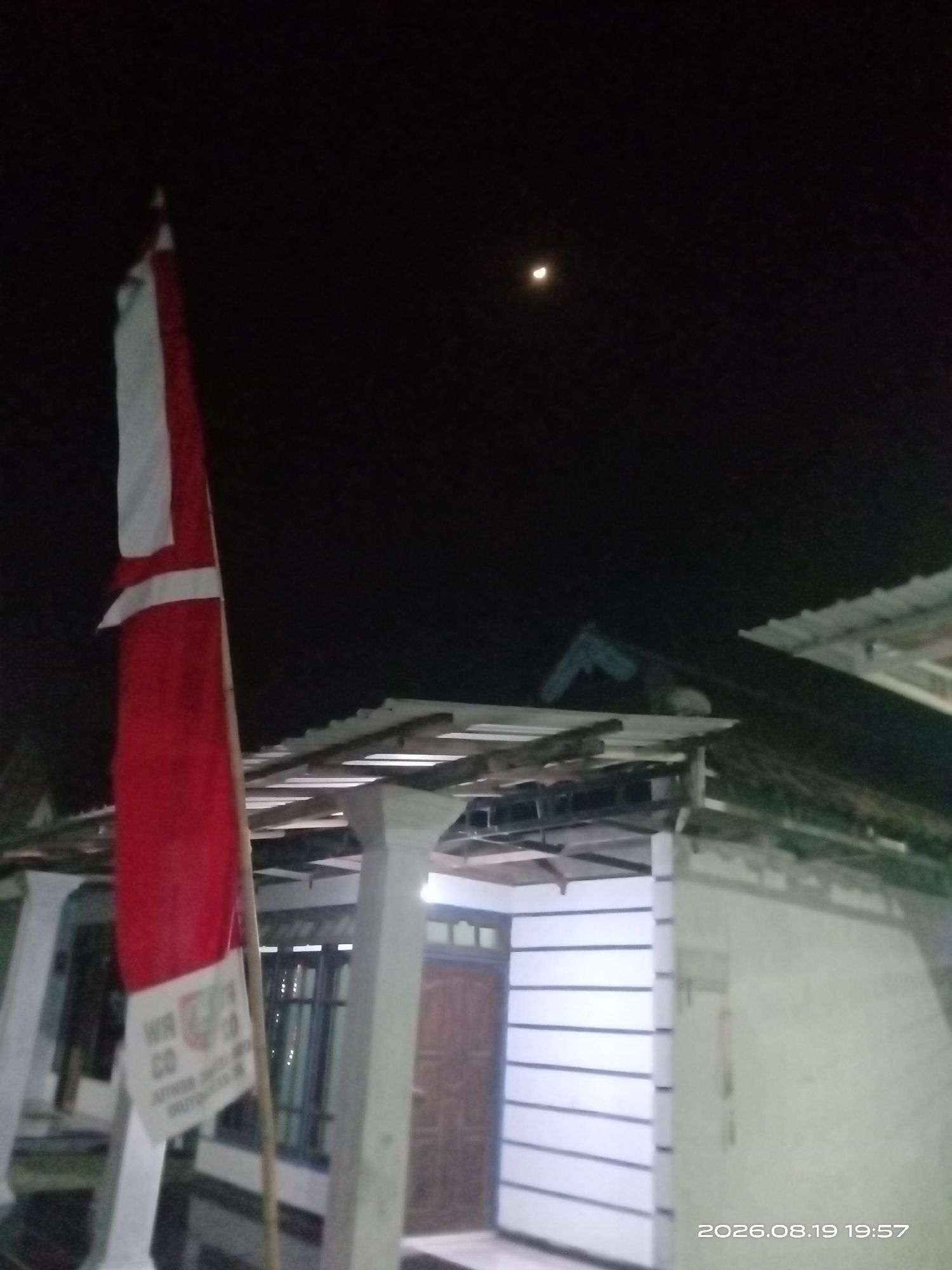 In Banyuarang, Indonesia, at night on August 19, 2026, at 19:57, a red and white flag is illuminated by a distant crescent moon in a dark sky. The flag is attached to a pole and hangs vertically. Behind the flag, a building with a white facade and a corrugated metal roof is visible. The building has windows and a wooden door. The scene appears to be an outdoor residential area.