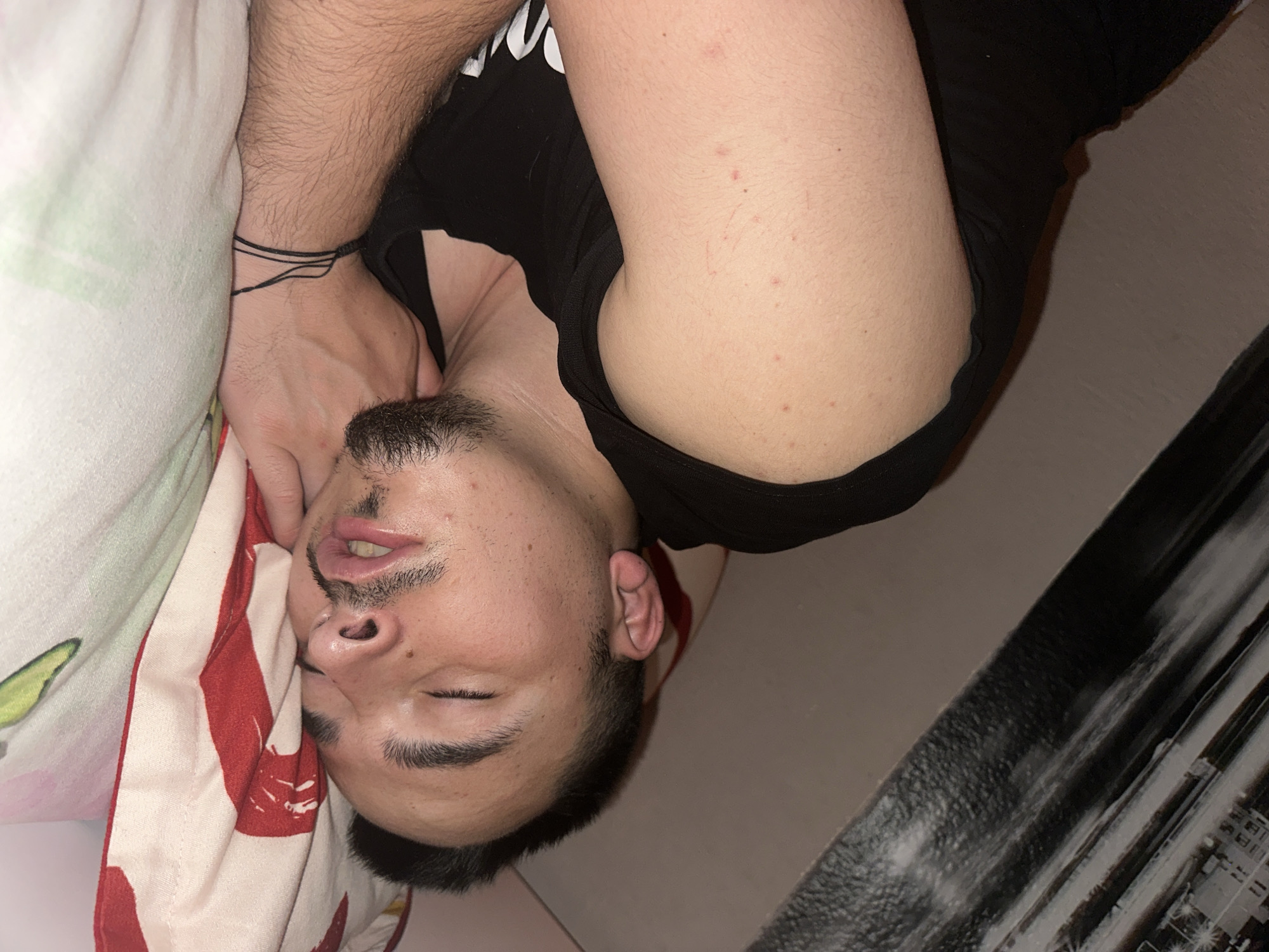 A man is asleep on a bed. The man has dark hair, a beard, and is wearing a black t-shirt. He is lying on a pillow with a red and white patterned pillowcase. His left arm is bent and his hand is near his face. The background includes a wall and a piece of furniture with a black and gray pattern. The scene does not seem to be in the city of Tulovo, Bulgaria.