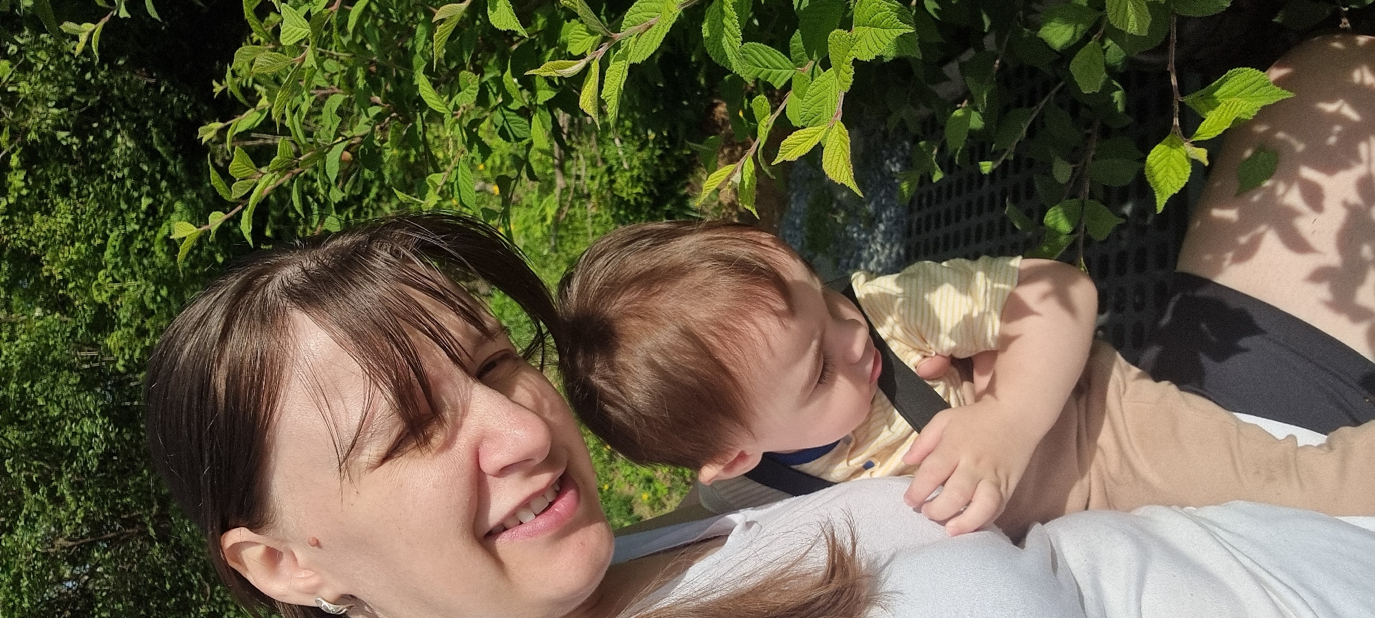 A selfie shows a woman and a baby outdoors in Dunnae-myeon, South Korea. The woman is smiling, with light skin and brown hair with bangs. She is wearing a white top and an earring is visible. A baby is nestled against her, wearing a yellow and white striped shirt and tan pants, with a dark strap across their chest. They appear to be reclining on someone’s lap. Green leaves fill the background, with dappled sunlight and shadows. A textured gray surface is also visible.