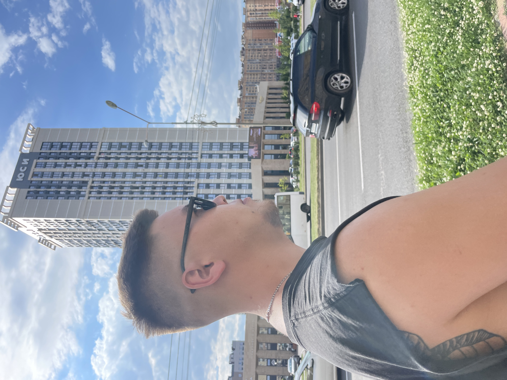 The image is a medium shot of a young man standing in front of a modern high-rise building. The man is wearing sunglasses and a dark t-shirt, and he is looking to his right. The building behind him is tall and slender, with many windows. The sky is blue with scattered clouds. In the foreground, there is a street with cars and a sidewalk lined with greenery. The overall scene suggests a day in a city. The building has the Cyrillic inscription "ЮСИ" on its upper facade. Based on the architecture and the presence of the "ЮСИ" inscription, which is associated with a construction company operating in Stavropol, Russia, the location is likely Stavropol.
