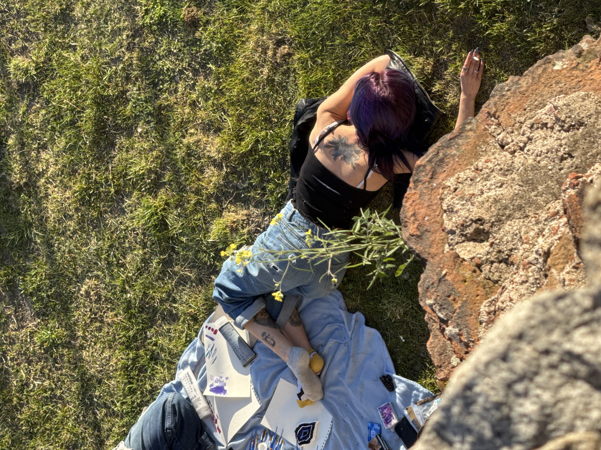 A high-angle shot depicts a woman lying on a grassy patch near a rock formation, potentially in Plovdiv, Bulgaria. The woman has purple hair, a tattoo on her back, and is wearing a black tank top and ripped jeans. She has long acrylic nails. She is partially lying on a blue blanket strewn with art supplies, including tubes of paint, brushes, papers with drawings and other small objects. The scene suggests an outdoor art session or picnic. The grassy area is sunlit, contrasting with the shadowed side of the rock formation.