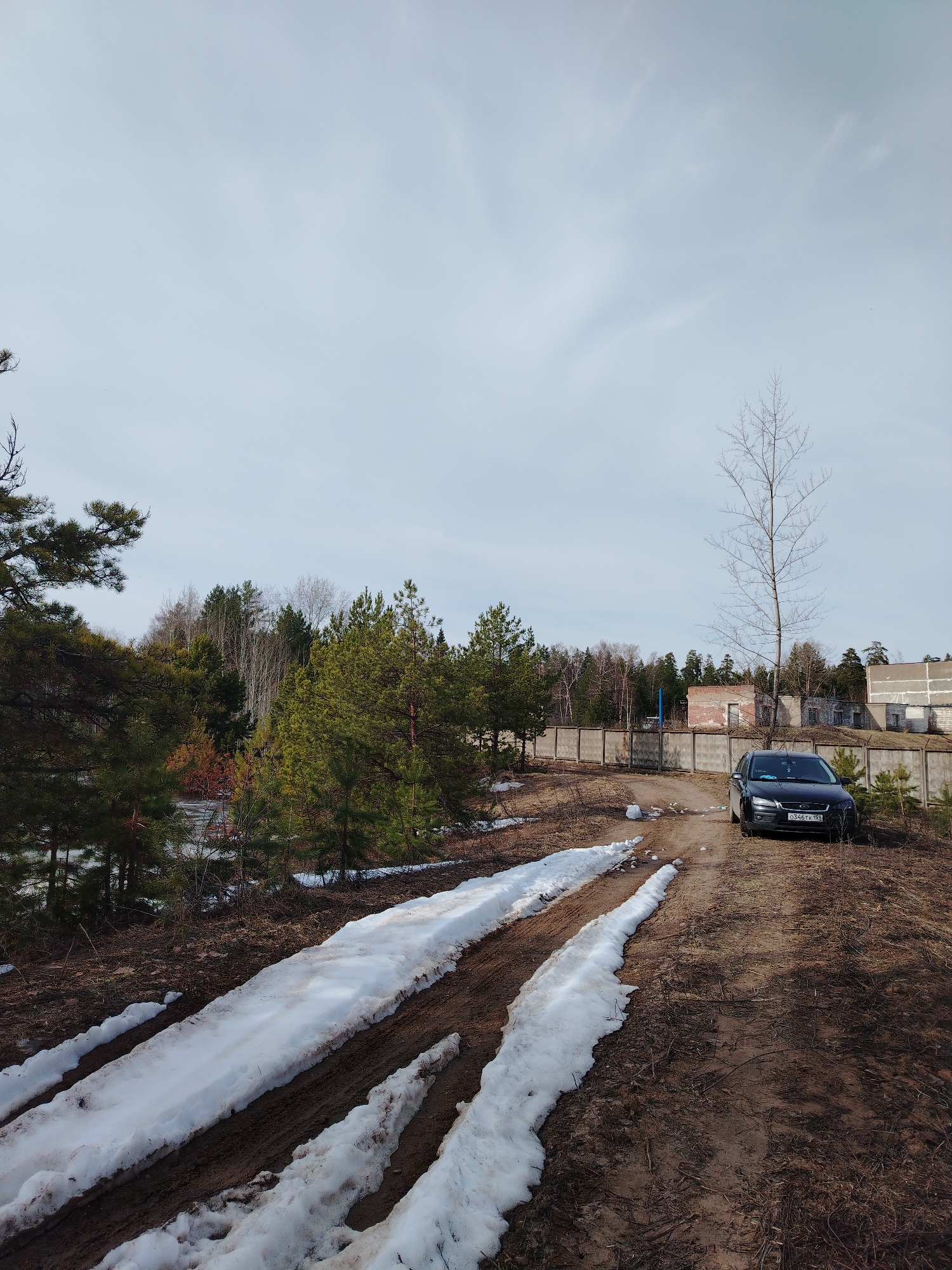 The image depicts a dirt road leading to a black vehicle parked near a concrete fence and derelict buildings. Patches of snow remain on the road's surface. The surroundings consist of pine trees and bare deciduous trees. The license plate on the car reads "О 346 ТК 152", indicating the car is registered in Nizhny Novgorod Oblast, Russia. The scene suggests a rural or semi-rural area, with the presence of abandoned or underutilized structures. Considering the provided information, the location is likely within Noviy, Russia, near an area with derelict buildings and natural woodland.