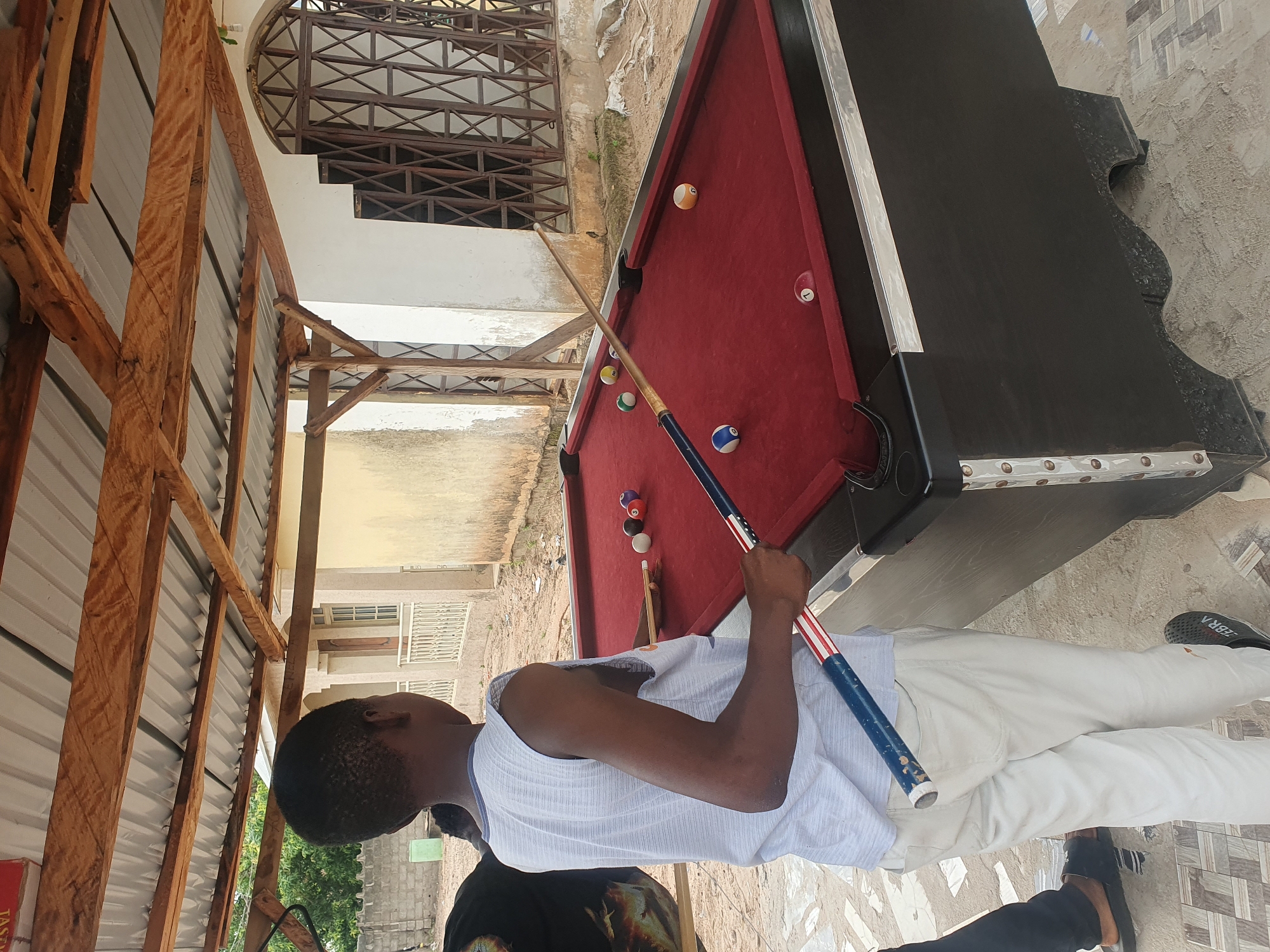 A young Black male, wearing a white sleeveless shirt and light-colored pants, stands on a tiled and dirt surface next to a red pool table. He holds a pool cue with an American flag design. Several pool balls are scattered on the table, including a white cue ball, a yellow ball, a blue ball, and a ball with the number 1. The pool table is situated outdoors under a wooden roof supported by wooden beams, with a corrugated metal roof above. In the background, a building with a barred window and white walls is visible. The environment appears to be in Lokoja, Nigeria, given the arid ground and the architectural style.