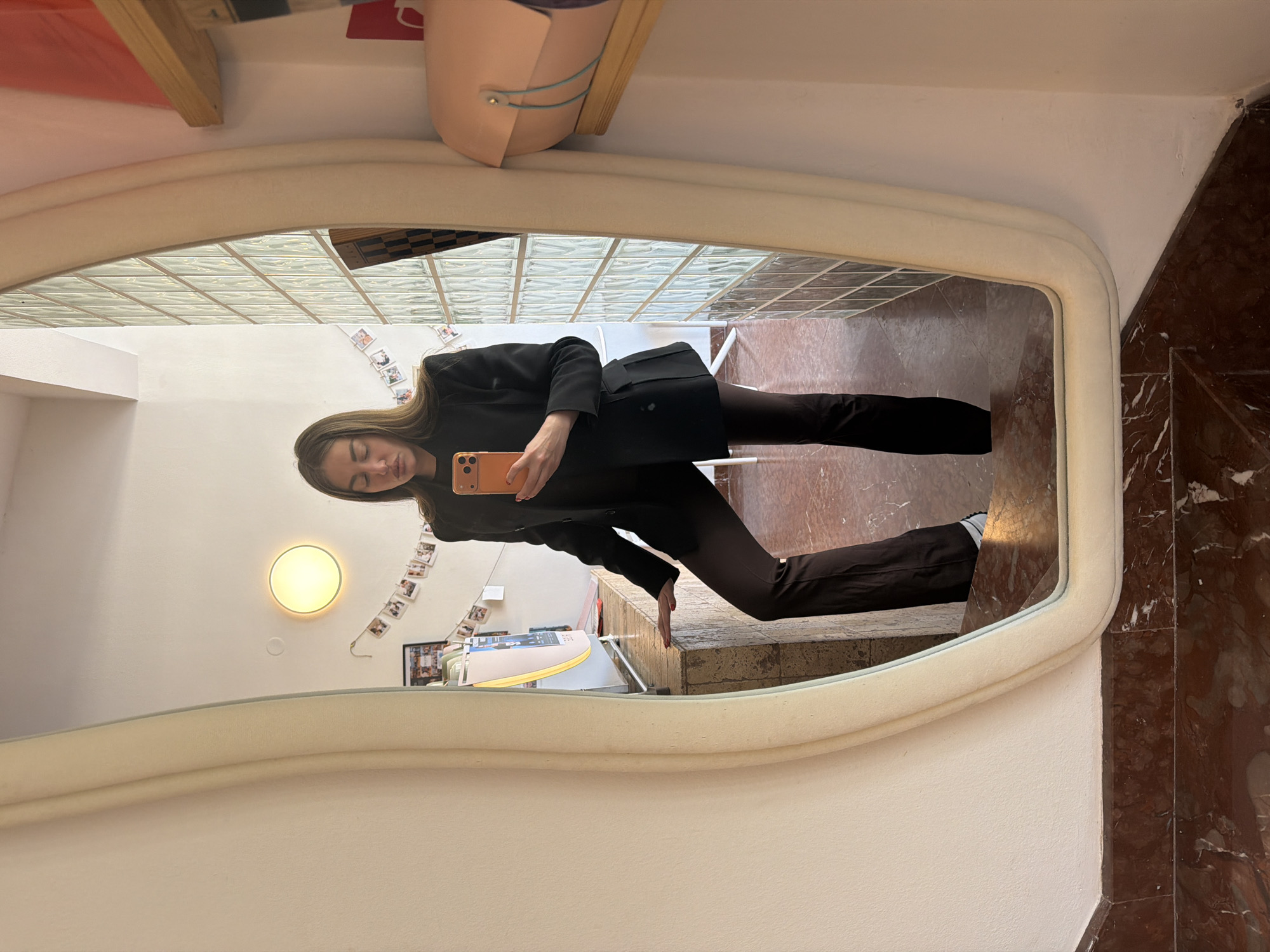 A young woman is captured in a mirror selfie in an indoor setting in Bar, Montenegro. The mirror, featuring an irregularly shaped, off-white frame, reflects her full body. She is holding an orange/peach-colored smartphone up to take the photo, with her head slightly tilted and her gaze directed towards the phone. She is dressed in a black blazer or jacket, dark brown or black trousers, and light-colored shoes with visible white socks. Her right hand, holding the phone, shows red nail polish.
The reflected background reveals a well-lit space. Behind the woman's reflection, there is a white wall with a circular, yellow-glowing light fixture mounted on it. Below the light, a string of small instant photos or polaroids is hung on the wall, adding a personal touch to the decor. Further back in the reflection, a glass block wall or window is visible, suggesting a source of natural light or an architectural feature. The original image's context (the part outside the mirror) shows brown marble flooring and steps, indicating a multi-level area or an entrance. A wooden shelf and a light-colored bag are also partially visible above the mirror frame. The scene appears to be daytime due to the bright, even lighting. No discernible text is visible.