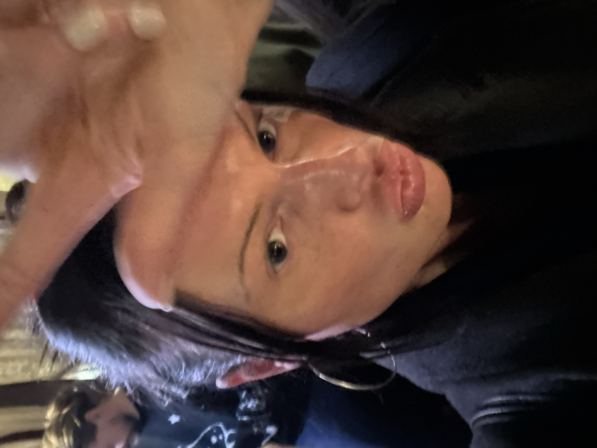 A close-up, rotated selfie shows a fair-skinned woman with dark hair, large eyes, and visible lip filler. She wears a black hoodie. Her hand is positioned close to the camera, obscuring part of her face. An earring is visible. In the background, another person is partially visible. The location cannot be determined.