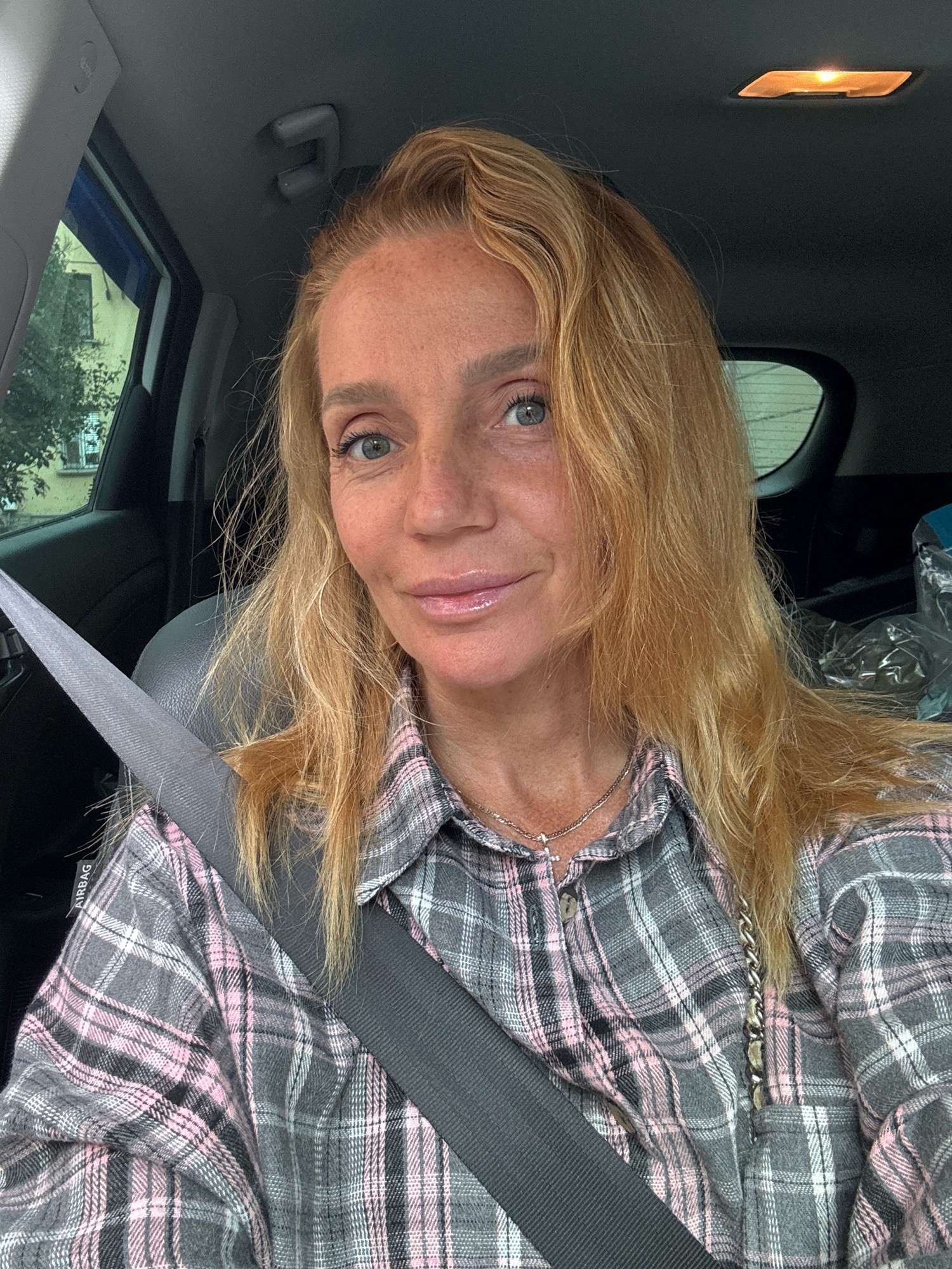 A woman with blonde hair and blue eyes, wearing a grey and pink plaid shirt and a seatbelt, is in the driver's seat of a car. She is looking at the camera and smiling. The interior of the car is visible, including the dashboard, steering wheel, and a window with a view of a building and trees. The scene appears to be in Saint Petersburg, Russia, based on the architecture visible through the window. The image is a selfie, taken from the perspective of the driver.