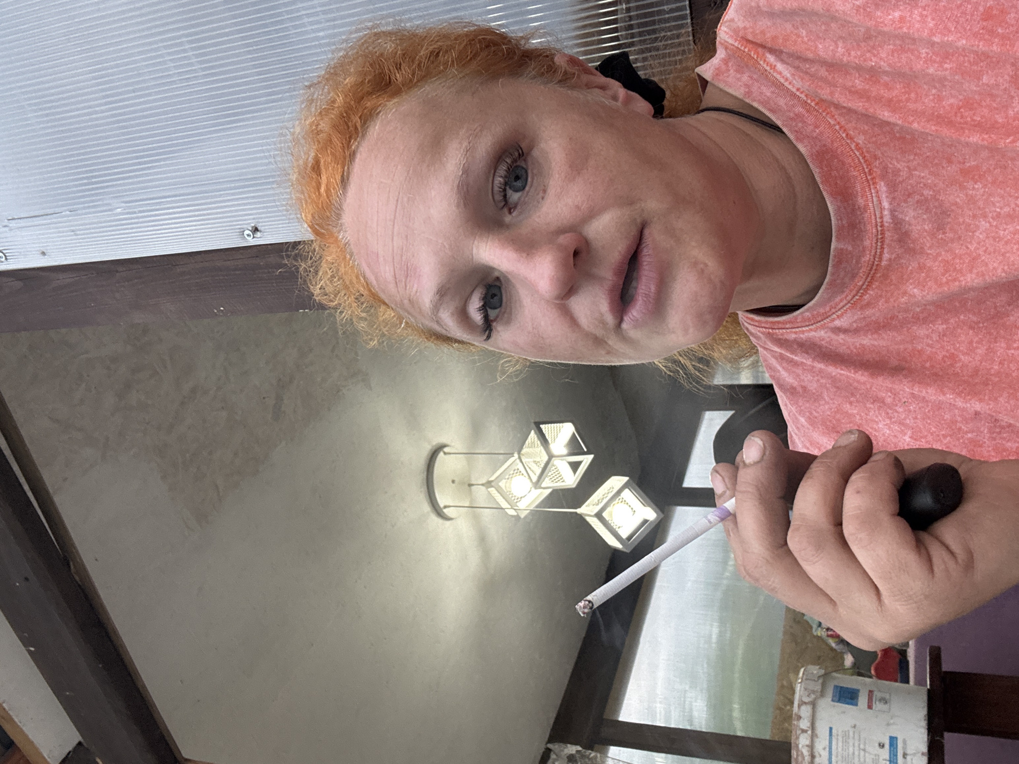 A woman with reddish hair, wearing a pink t-shirt, is holding a lit cigarette in her left hand and a small black object in her right hand. She appears to be indoors, with a ceiling of corrugated metal panels and wooden beams visible above her. A light fixture with three geometric lamps hangs from the ceiling. The photograph was taken indoors in the city of Chaikovsky, Russia.