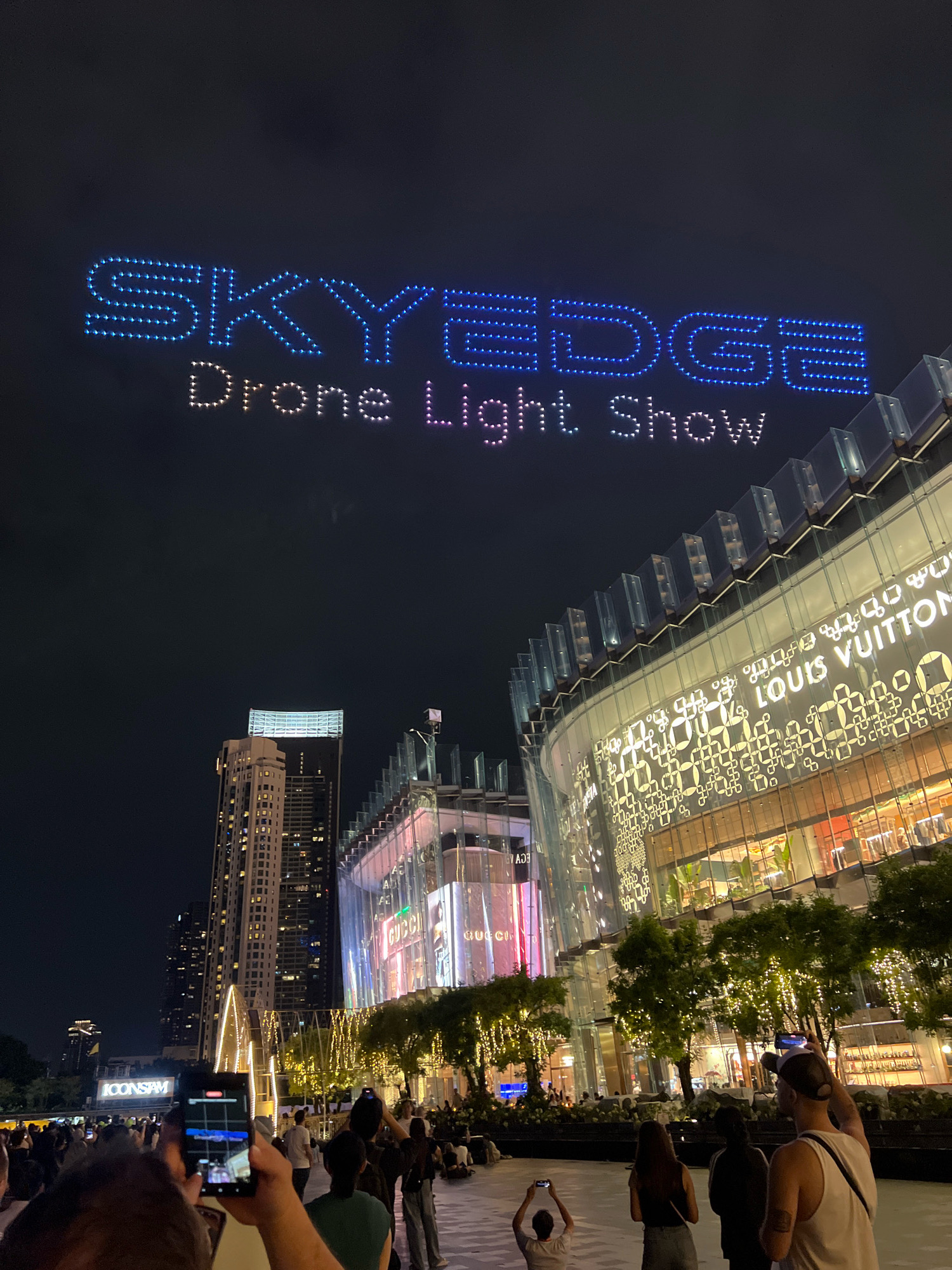 An aerial view of a drone light show is depicted against a night sky, with the words "SKYEDGE" and "Drone Light Show" formed by illuminated dots. Below, the ICONSIAM shopping mall is visible in Bangkok, Thailand, with its distinctive architecture and illuminated facade displaying brands like Louis Vuitton and Gucci. A crowd of people are gathered, many holding up their mobile phones to capture the event. The environment is urban, characterized by tall buildings and festive lighting.