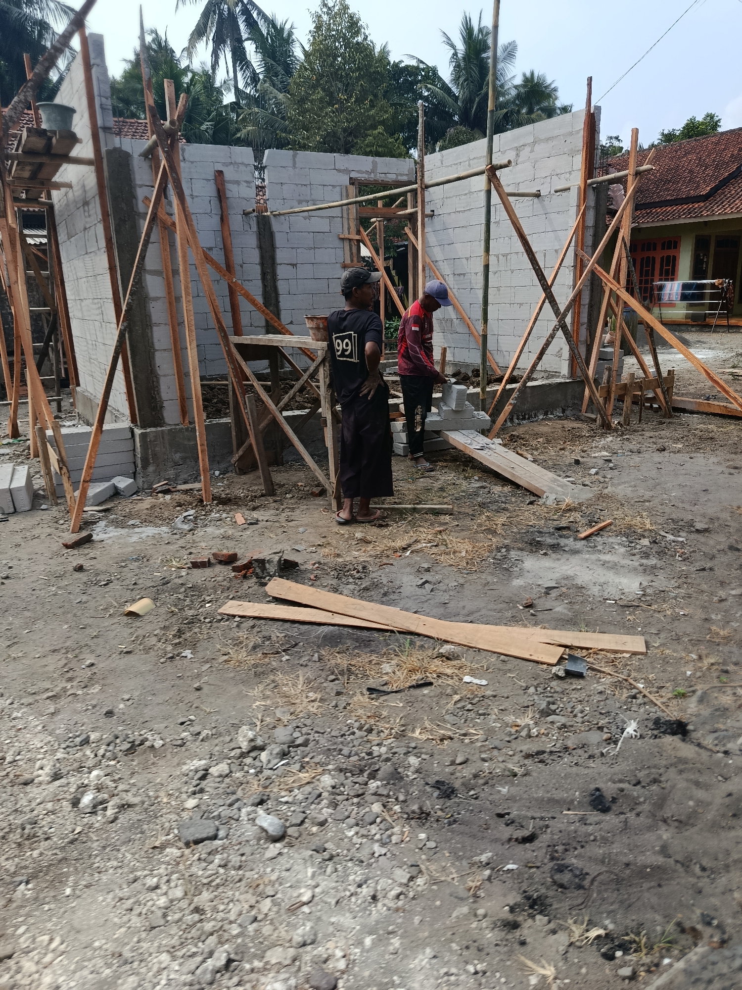 In Desa Rawajaya, Indonesia, a construction site is depicted. Two men are actively engaged in building work. One man, wearing a black t-shirt with the number "99" on it and dark pants, stands near the foreground, observing the work. The second man, dressed in a red and black long-sleeved shirt and a blue cap, is positioned further back, placing cinder blocks. They are working on a structure made of concrete blocks, supported by wooden scaffolding and bamboo poles. The ground is covered in dirt, gravel, and scattered construction materials like wooden planks and bricks. In the background, a residential building with a red-tiled roof is visible, along with palm trees and other foliage, suggesting a semi-rural or suburban environment. The sky is overcast.