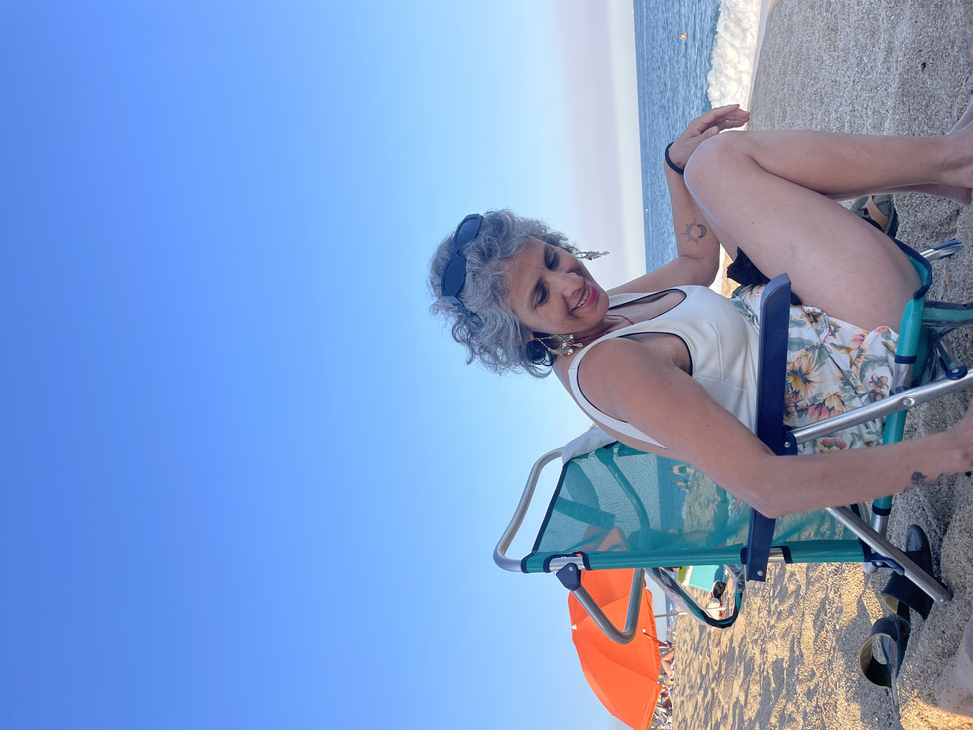 A woman with gray curly hair and sunglasses on her head sits in a teal beach chair on a sandy beach. She is wearing a white tank top and floral shorts. She is looking down and smiling. The ocean is visible in the background with waves breaking on the shore. An orange umbrella is set up on the beach to the left of the woman. The sky is clear and blue. This scene is taking place on the beach in the city of Canet de Mar, Spain.