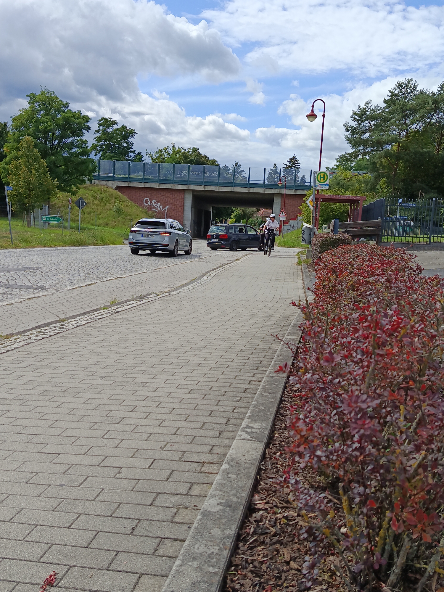 The image shows a street scene in Groß Köris, Germany. A paved path, likely a sidewalk or bike lane, occupies the foreground and extends into the distance. To the right, a dense bush with red-tinged leaves borders the path. Beyond the path, a road is visible with a silver station wagon and a dark sedan driving away. A cyclist wearing a white helmet and a white shirt is also moving away from the camera on the paved path. In the background, a bridge crosses over the road, with a bus stop shelter and a lamppost visible near the bridge. Green trees and a grassy incline are present on either side of the road and under the bridge. The sky is partly cloudy. A street sign with a green arrow pointing left indicates "Schul- und Wohngebiet". Another sign with a green circle and an 'H' signifies a bus stop. Graffiti can be seen on the brickwork of the bridge.