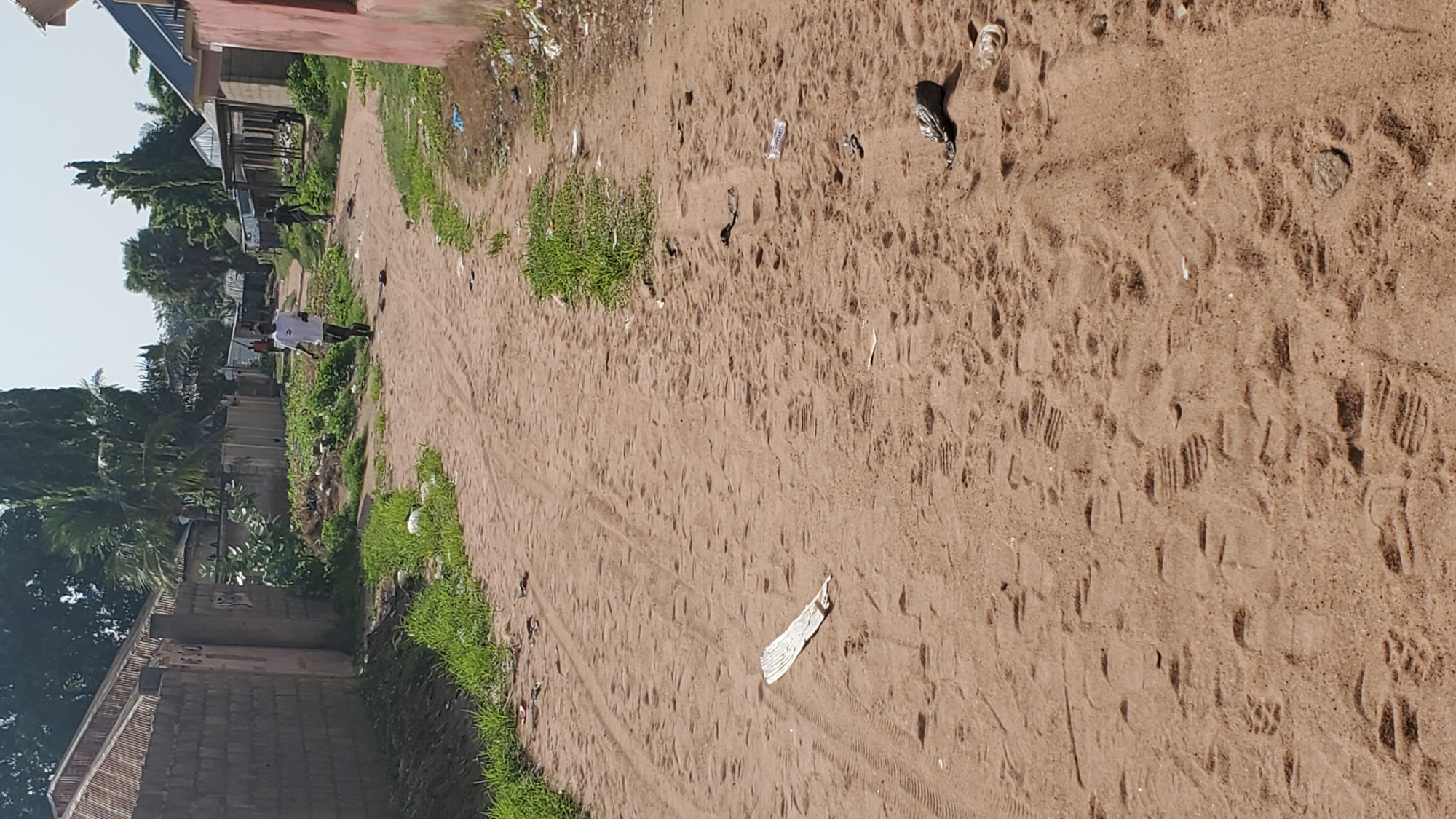 The image depicts a sandy street in Jalingo, Nigeria. The road is unpaved and shows tire tracks and numerous footprints in the sand. On the left side of the street, a brick wall lined with sparse greenery borders the path. On the right, more buildings are visible, some appearing to be shops or houses with corrugated iron roofs.
In the mid-ground, a young man wearing a white t-shirt and dark shorts is walking away from the camera. Further in the distance, another individual is walking in the same direction. The environment appears to be a residential or commercial area within the city, with several trees and vegetation adding to the scene. Litter, including plastic bottles and bags, is scattered along the sides of the road, indicating the presence of waste. The overall impression is a typical street scene in a Nigerian city, characterized by its sandy terrain and modest structures.