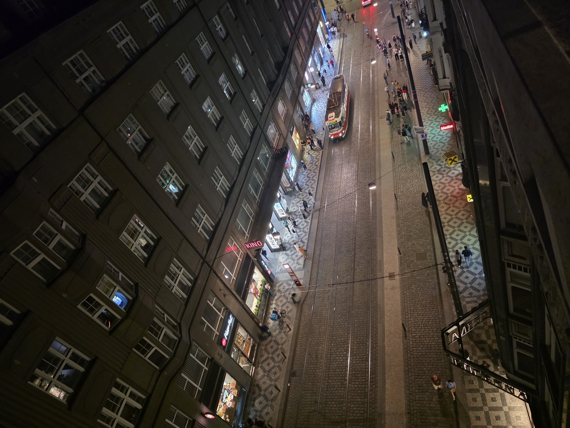The image depicts an aerial view of a street at night in an urban setting. A tall building with numerous windows stands on the left side of the frame. A tram runs down a street with tram tracks. Pedestrians walk along the sidewalks and cross the street. Storefronts are visible along the side of the building. Streetlights illuminate the area. The street is paved with cobblestones.