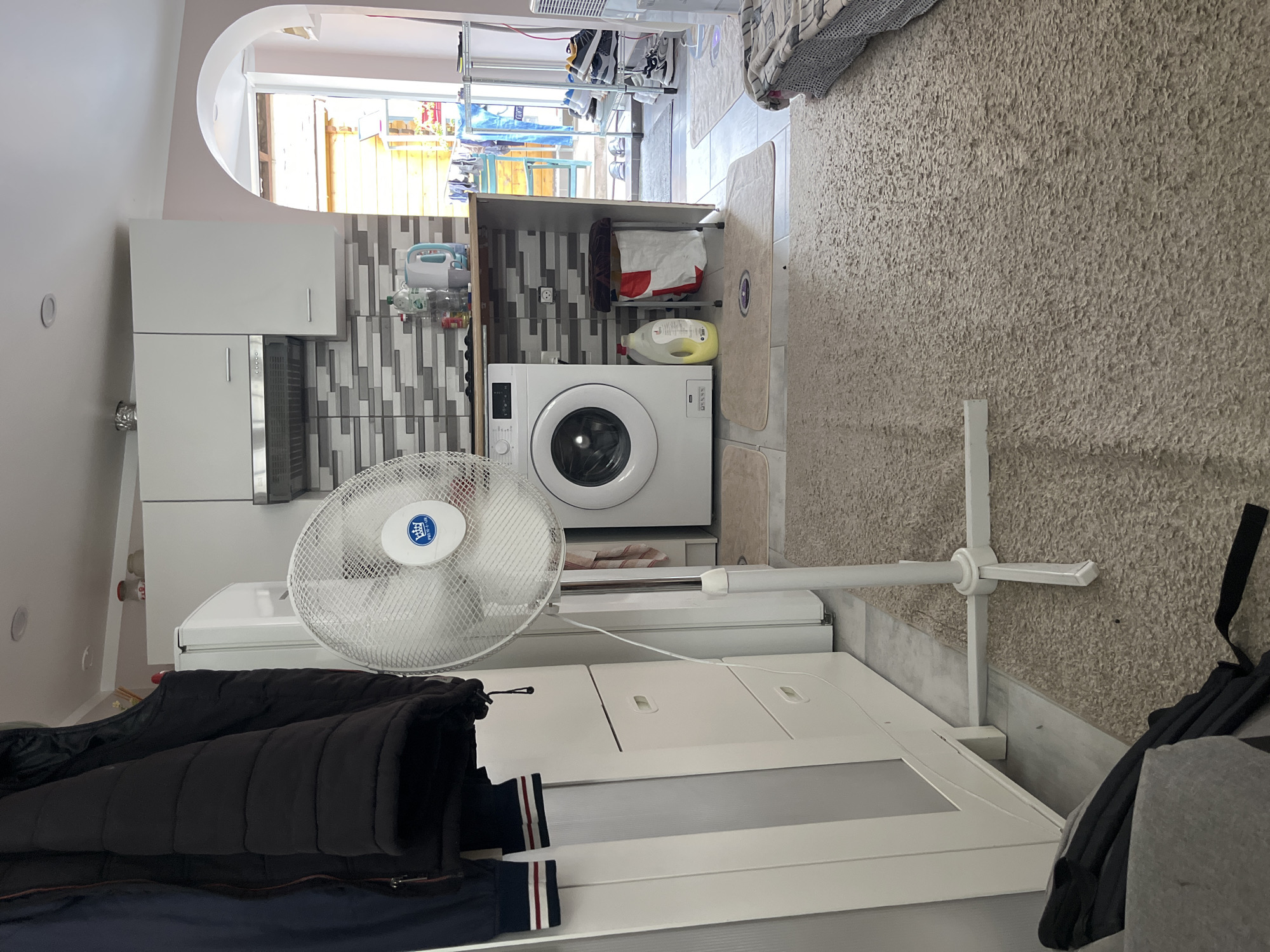 The image depicts an indoor scene, likely a laundry area or kitchen adjacent to a living space, located in the city of Stains, France. A white standing fan is positioned in the foreground, facing the viewer. To its left, a tall, white cabinet or appliance, possibly a refrigerator or wardrobe, is visible, with two dark jackets hanging on its side. Behind the fan, a white front-loading washing machine is present. The background reveals a kitchen area with white cabinets, a tiled backsplash, and a countertop. Beyond the kitchen, an open doorway leads to an outdoor space with wooden structures and laundry on display. The floor is covered with a beige carpet in the foreground and tiled in the background. No people are visible in the scene.