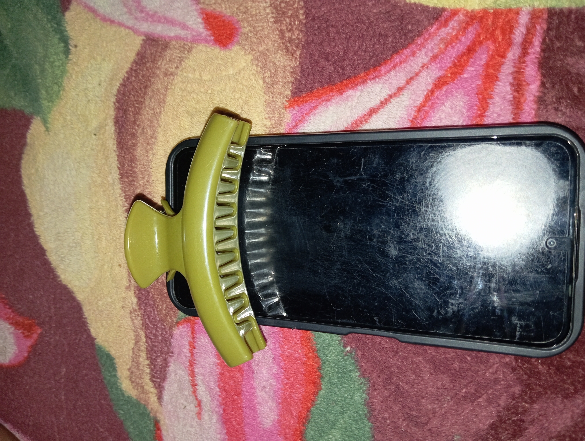 A green hair claw clip is placed on top of a black smartphone, which is lying on a multi-colored, fuzzy blanket. The blanket has abstract patterns in shades of pink, red, green, and yellow. The smartphone's screen is visible, showing numerous scratches and a bright reflection. The hair clip is positioned horizontally across the top of the phone, with its teeth facing downwards. The image is taken from a top-down perspective. There are no people or other objects visible in the frame. The scene is indoors, likely on a bed or sofa, given the texture of the blanket. The overall lighting suggests an indoor setting with artificial light contributing to the glare on the phone screen. The location of Rejosari, Indonesia is not discernible from the visual information provided in the image.