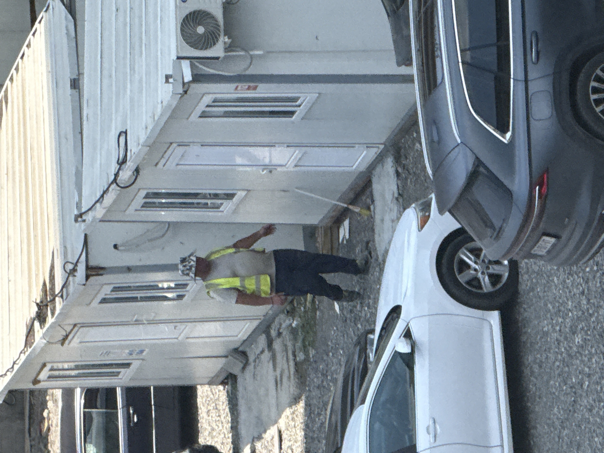 A man in a reflective vest and a patterned bucket hat walks next to a row of portable buildings. He is wearing a light-colored t-shirt and dark pants. The buildings appear to be modular units, possibly used as temporary offices or storage. A white car is parked in front of one of the buildings, and a dark-colored SUV is partially visible in the foreground. The ground is covered with gravel and some patchy dirt. An air conditioning unit is mounted on the side of one of the buildings. The overall scene suggests an outdoor, possibly construction or industrial area. The image was taken in Tbilisi, Georgia.