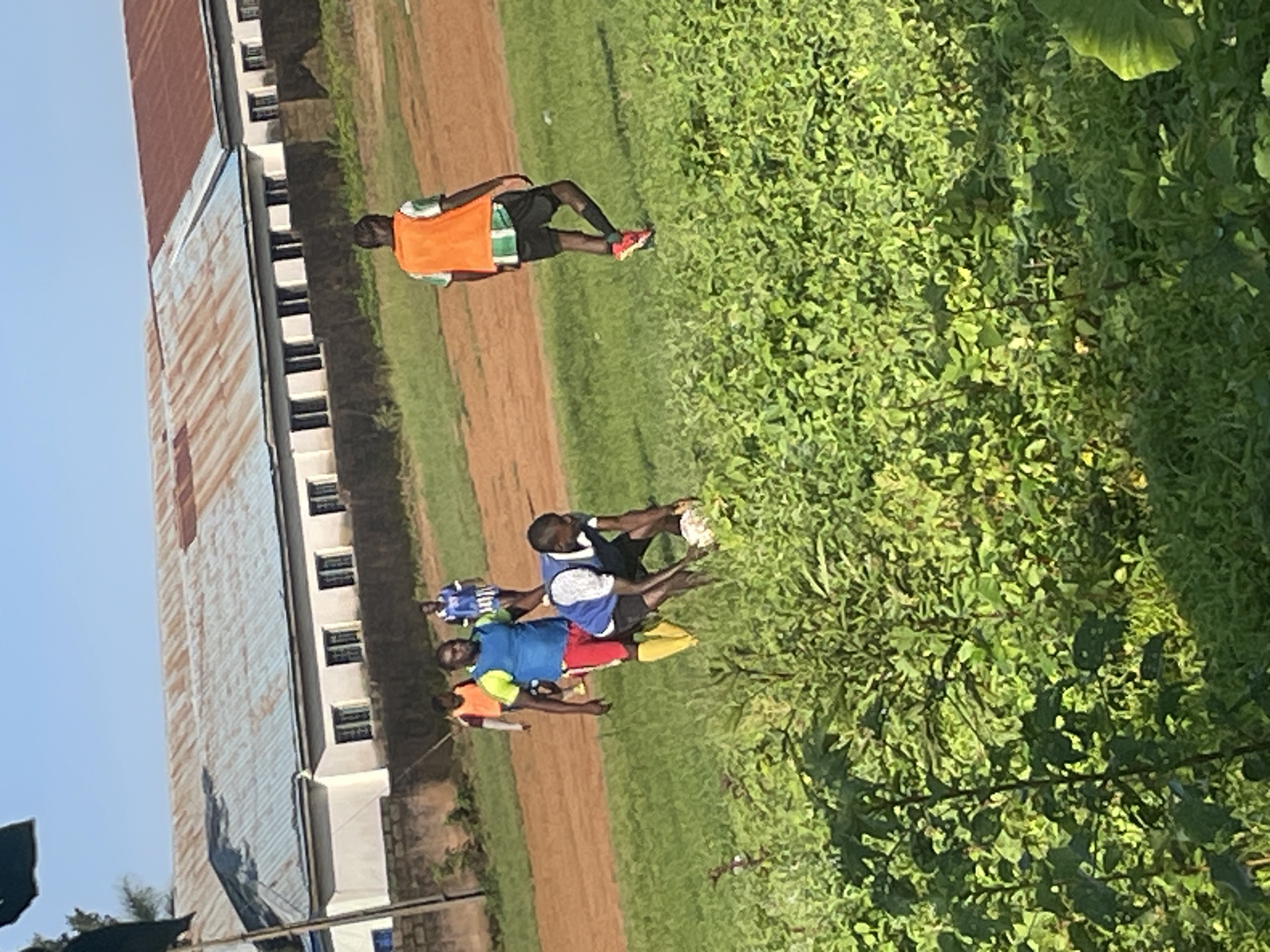 In Jos, Nigeria, on a dirt field adjacent to a building with a corrugated metal roof, a group of men are playing soccer. One man, wearing a blue shirt and red shorts, is bending down to pick up the soccer ball. He is surrounded by other men in athletic wear, some in orange vests and others in blue and white striped jerseys. The field is partially covered in grass and has a dirt track running alongside it. The building in the background appears to be a school or administrative building, with a concrete wall and barred windows. The sky is clear and blue.