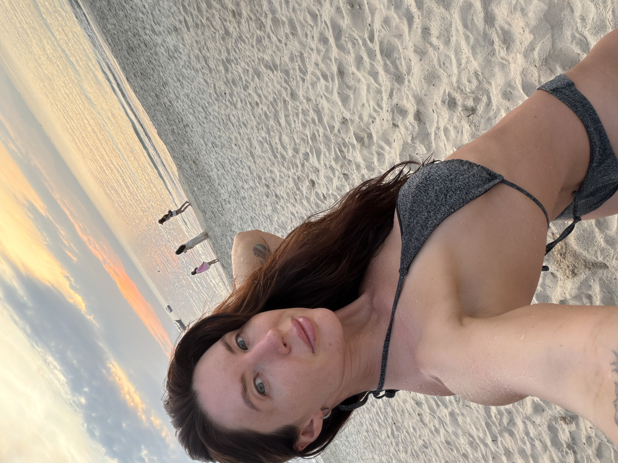 A woman with long, dark hair and fair skin, wearing a sparkling gray bikini, takes a selfie on a sandy beach at sunset. She has blue eyes, full lips, and light makeup. Her left arm is behind her head. In the background, the ocean is calm and reflects the warm hues of the sunset sky, with a few small boats visible on the horizon. Three other women are standing on the beach, farther away from the camera. The sand is light-colored and textured. The scene appears to be in Dong Ninh Hoa Ward, Vietnam, based on the provided context.