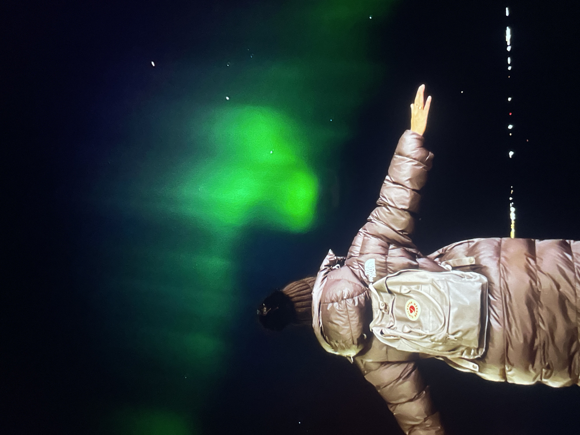 A figure in a brown puffer jacket and a beige Fjallraven backpack stands against a dark backdrop punctuated by a vibrant, green, aurora-like display. The figure's right arm is raised with an open hand. To the right of the figure, a vertical series of white and red lights descends, creating a visual contrast against the dark sky. The location is city Unknown, Unknown.