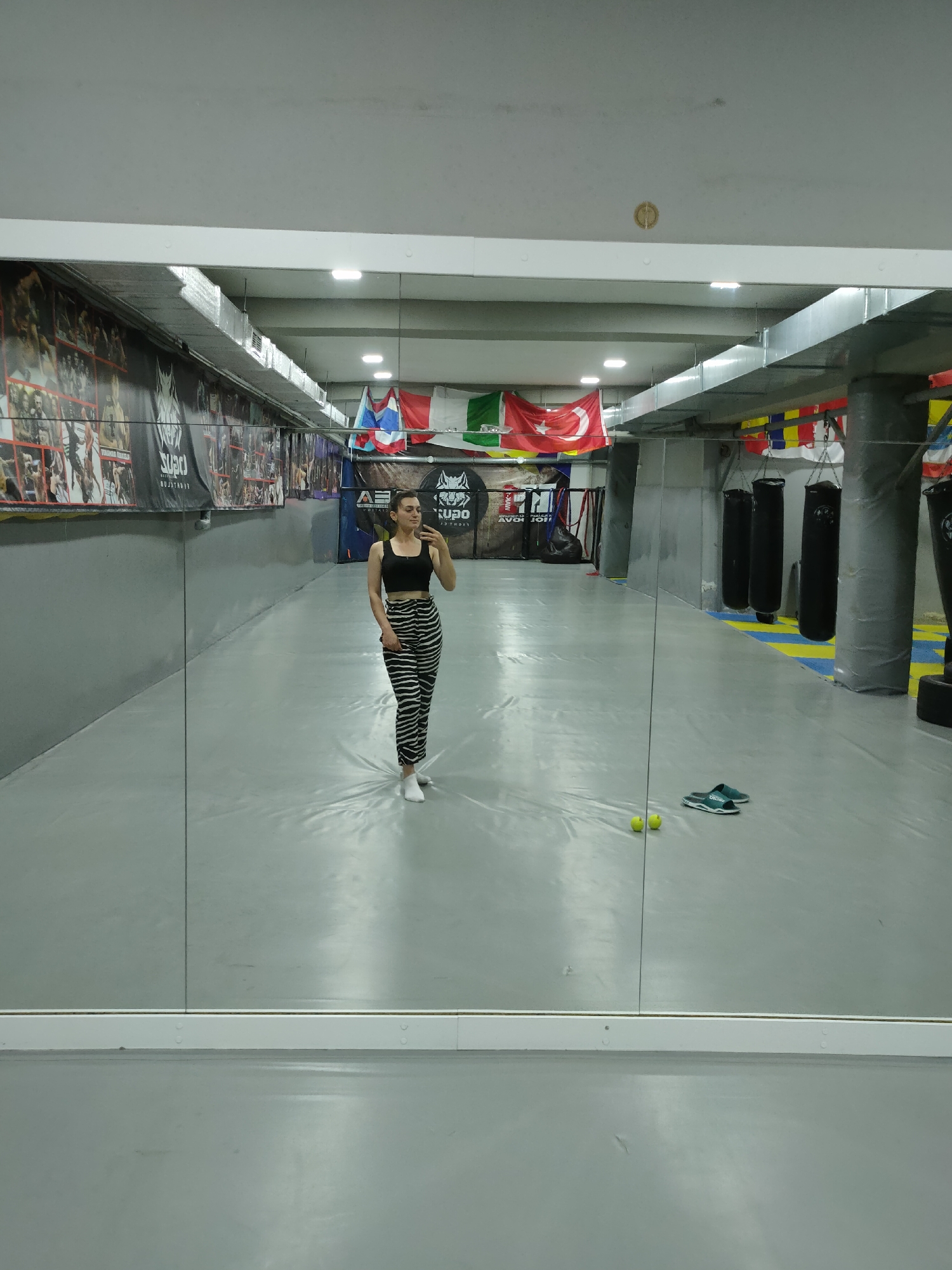 A woman in a black tank top and black and white striped pants stands in front of a full-length mirror in what appears to be a gym or martial arts studio. She is holding a phone in her right hand, taking a selfie. The studio has a grey floor and walls, with large posters and flags hanging on the walls. Several punching bags are visible in the background. Two yellow tennis balls and a pair of teal slip-on shoes are on the floor in front of the mirror. The woman is in the city of Comrat, Moldova.