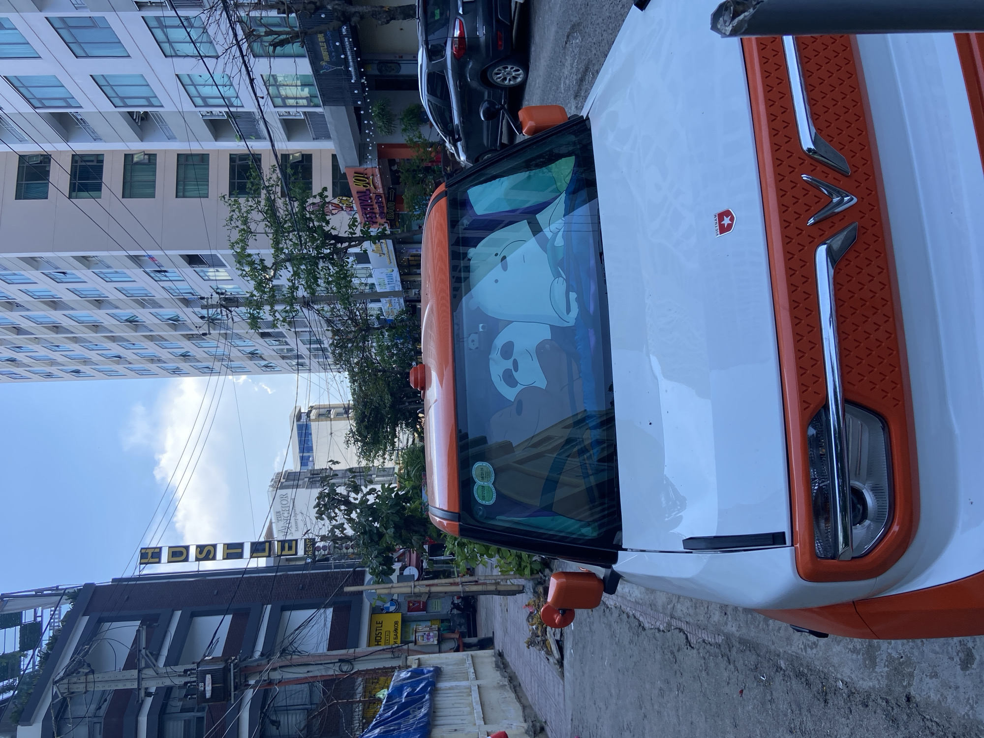 In Bac Nha Trang Ward, Vietnam, a white and orange electric car with cartoon bears displayed on its windshield is parked on a paved street. The car faces forward, with its headlights visible. The street is lined with buildings of varying heights, including a tall apartment building and a multi-story building with shops on the ground floor. Electrical wires are strung overhead. A dark-colored SUV is parked behind the electric car. The ground is paved with concrete and has some debris. The sky is partly cloudy.