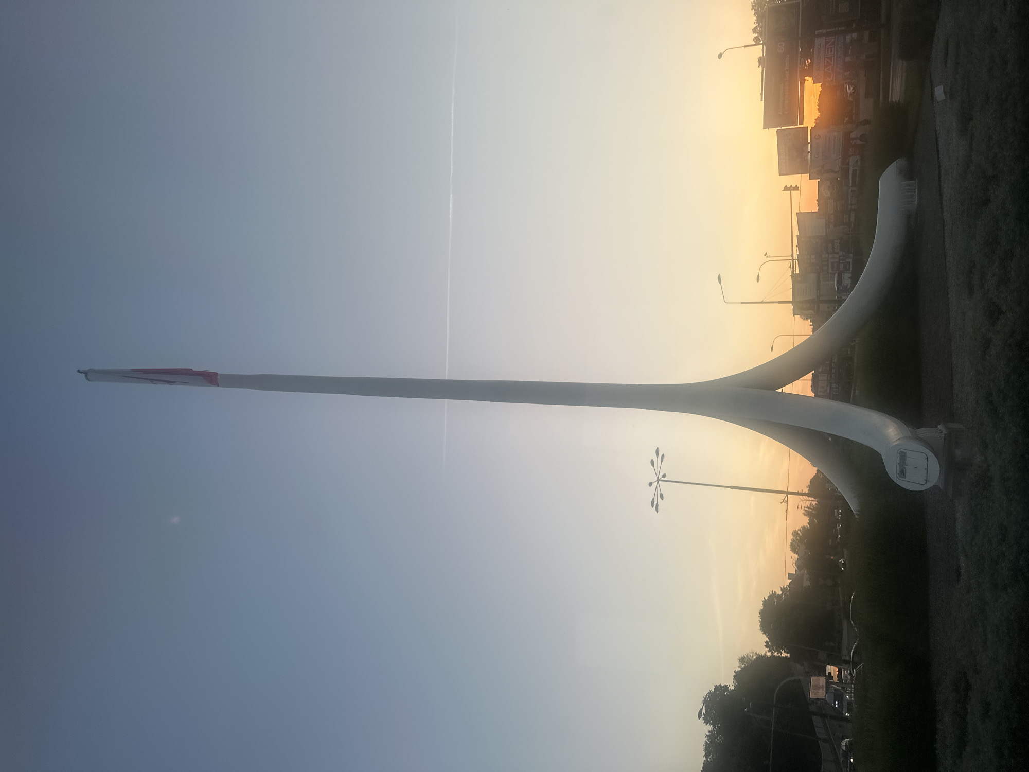 The image depicts a large, abstract white sculpture with three curving legs that meet at the base, supporting a tall, slender pole topped with a flag. The sculpture is situated in a grassy area, likely a roundabout or public square, with a road visible in the background. Several streetlights, some with multiple lamps, are scattered throughout the scene. The sky is clear and shows a gradient from a light blue at the top to a warm orange-yellow near the horizon, indicating it is either sunrise or sunset. A faint contrail is visible in the sky. In the background, buildings and billboards are discernible, suggesting an urban setting. Traffic lights and vehicles are also present on the road. The overall impression is that of an outdoor public space in an urban environment during twilight. This sculpture is the "Biała Dama" (White Lady) located at Plac Dworcowy in Lublin, Poland.