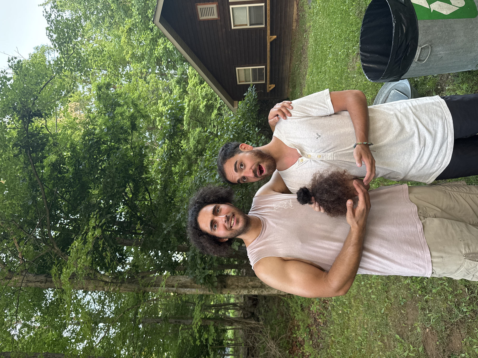 Two men are standing in front of a wooden cabin with a grassy yard and trees in the background. The man on the left, with curly dark hair and a beard, is wearing a light pink sleeveless t-shirt and khaki shorts. He is holding a clump of dark, curly hair in his hands. The man on the right, with short dark hair and a beard, is wearing a light grey t-shirt and dark shorts. He has a surprised expression on his face and his left arm is around the man on the left's shoulders. A metal trash can with a black liner and a recycling symbol is to the right of the men. The scene appears to be outdoors in a wooded area. The image does not provide any specific information about the city of Washington Township, United States of America.