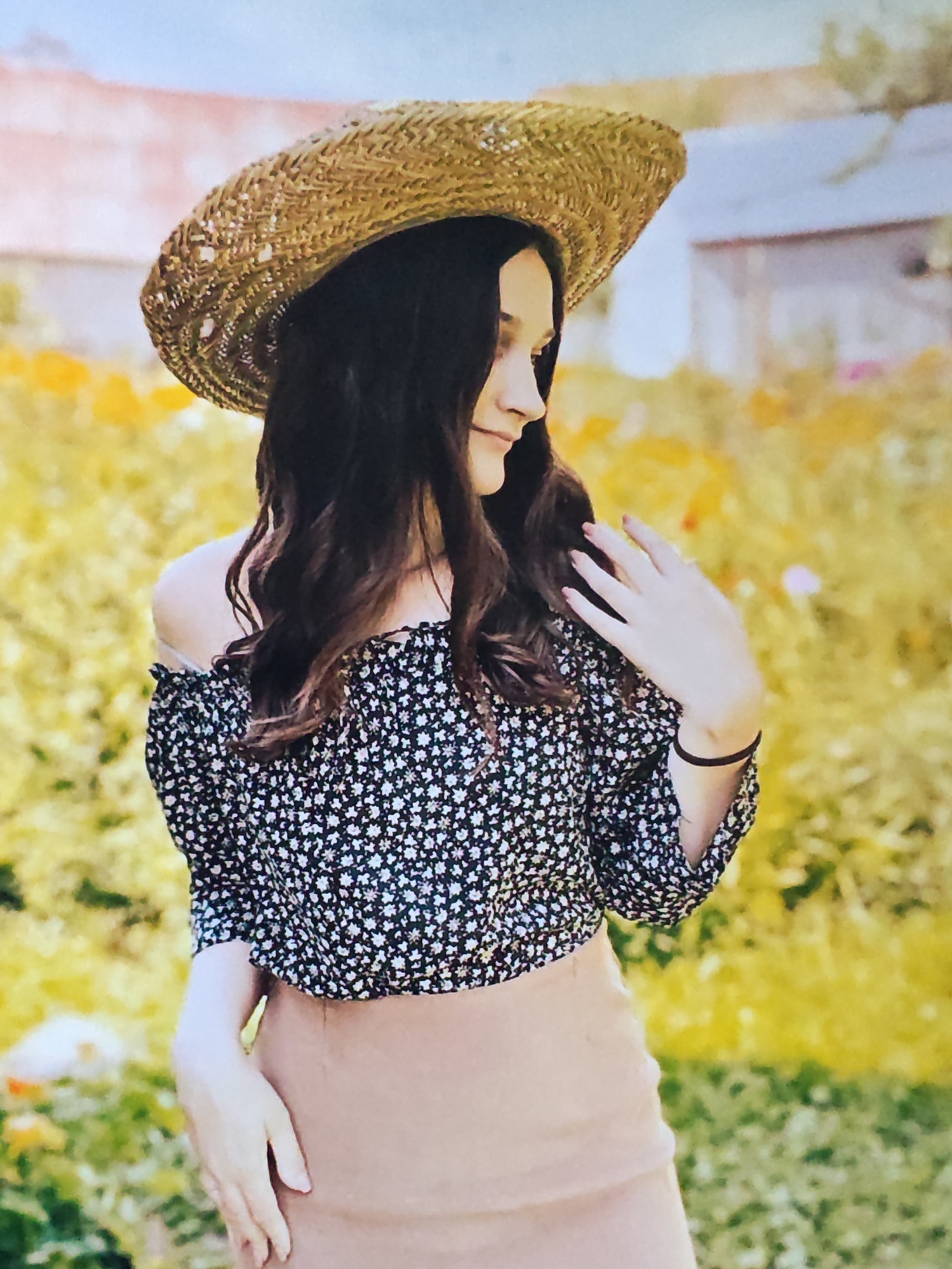A young woman stands in a field of yellow flowers, wearing a straw hat and a floral off-the-shoulder top. She has long, dark, wavy hair and is looking to the side with her right hand gently touching her hair. She is wearing a light brown skirt. The background is blurred, showing more of the yellow flowers and some muted colors that suggest buildings or structures in the distance.