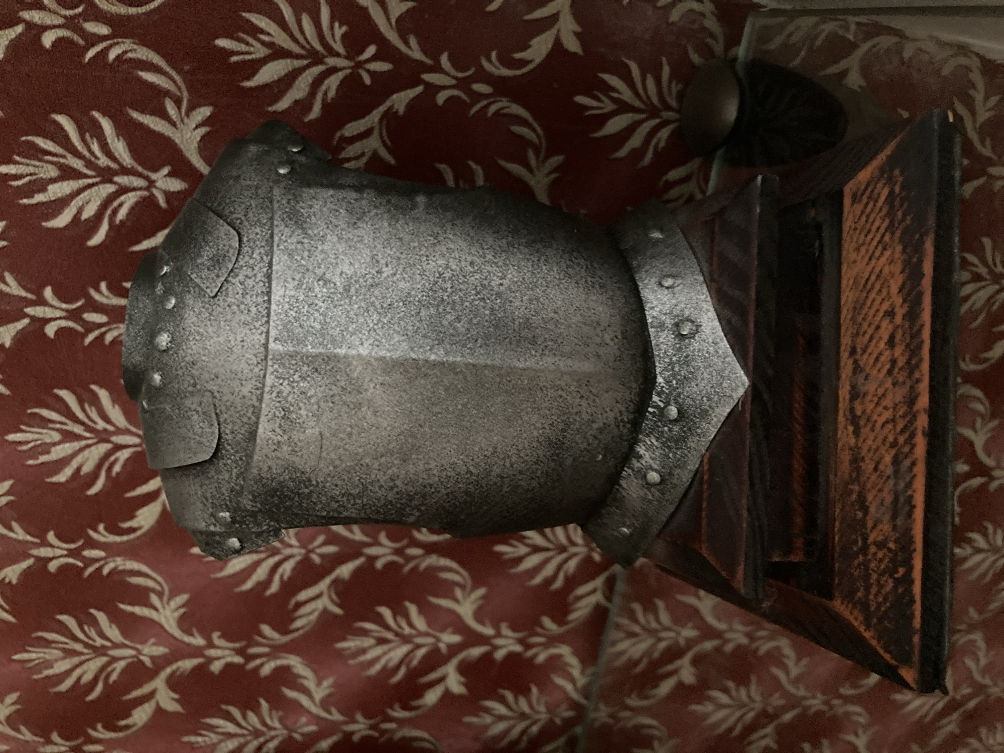 A half-torso, metallic suit of armor, possibly a decorative replica, is displayed on a wooden pedestal against a red damask-patterned fabric backdrop. The armor is a dull silver color with rivets visible along its edges, particularly around the shoulder and neck areas. The pedestal is multi-tiered, made of dark wood with visible grain and a reddish-brown hue. To the right of the armor, a portion of a dark, rounded object, possibly a helmet or a decorative finial, is visible. The background fabric exhibits a repeating pattern of stylized light-colored floral or leaf motifs. The lighting is diffused, casting subtle shadows. The scene is devoid of any human presence.