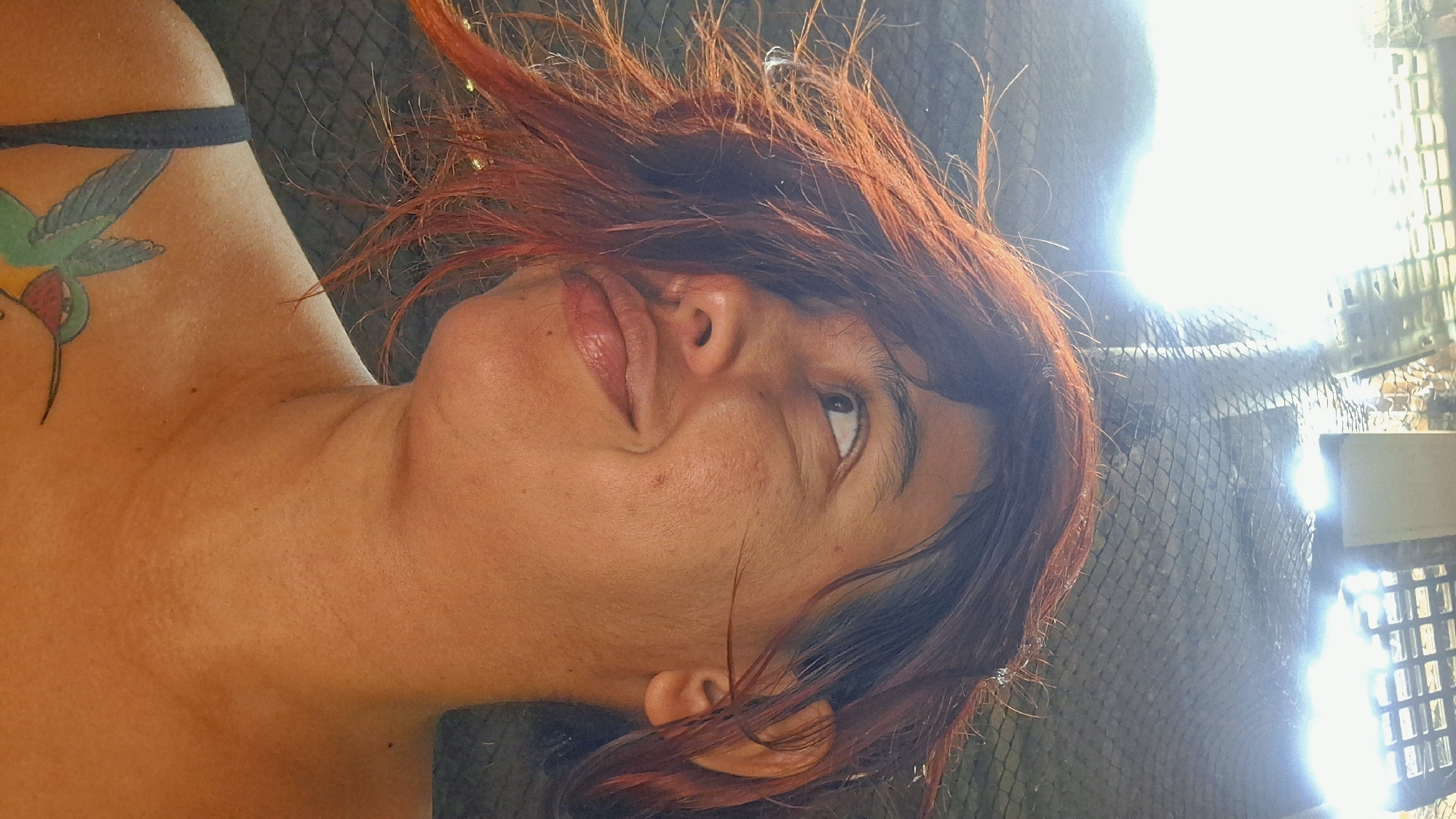 A woman with reddish-brown hair and a tattoo of a hummingbird on her left shoulder is pictured in what appears to be an outdoor setting in Vallecito, Venezuela. She is looking up and to her right with a slight smile. The background is out of focus but suggests a structure with a woven material and bright lights. The overall lighting is harsh, creating strong highlights and shadows on the woman's face and skin. The image is a close-up, focusing on the woman's upper body and face.