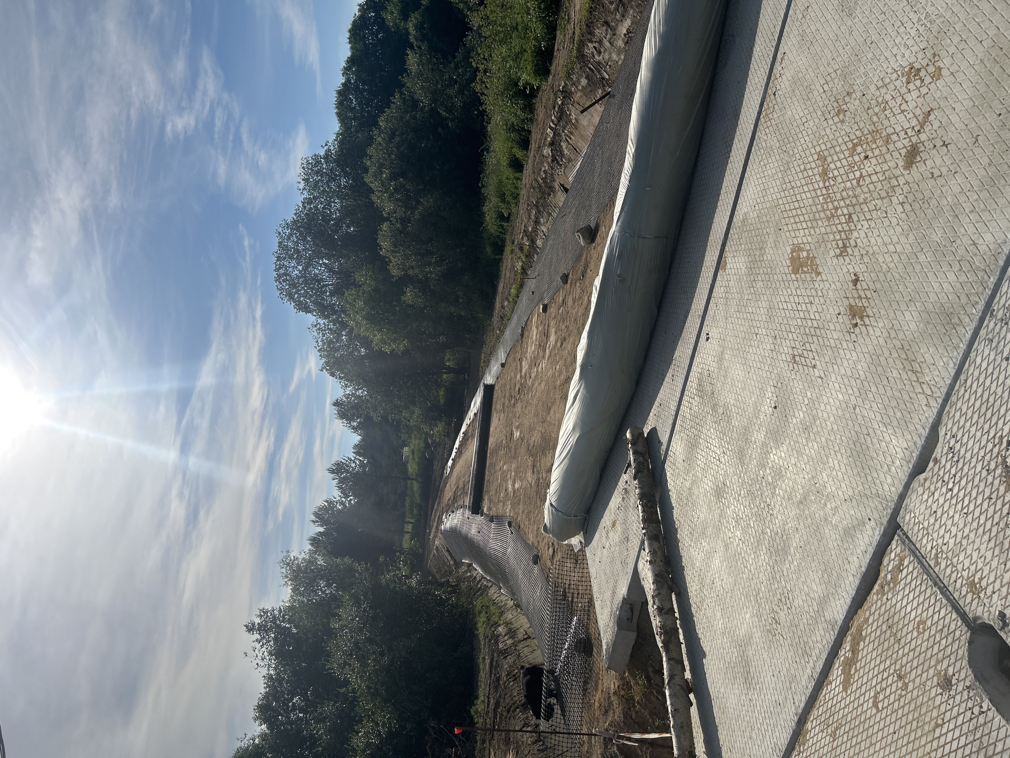 This image depicts a construction site, likely in Svetloe, Russia, with a focus on erosion control or slope stabilization. The scene is outdoors, under a partly cloudy sky with bright sunlight. In the foreground, several large, diamond-patterned concrete slabs are laid out, suggesting a temporary pathway or work platform. A large, rolled-up grey tarp or geotextile material lies on these slabs, positioned along the right side of the frame.
The middle ground reveals a sloped area being prepared for construction. A mesh or geotextile fabric, possibly for soil reinforcement or drainage, is spread across the slope and down into a trench-like excavation. The soil appears to be a mix of sand and gravel. Several large stones are visible on the fabric. Along the left side of the slope, a black mesh netting is installed, possibly to prevent debris from falling. A red marker is planted in the ground near the netting.
The background is dominated by a dense line of lush green trees, forming a natural backdrop to the construction activity. The overall impression is one of ongoing earthwork and infrastructure development in a natural environment. There are no people visible in the image.