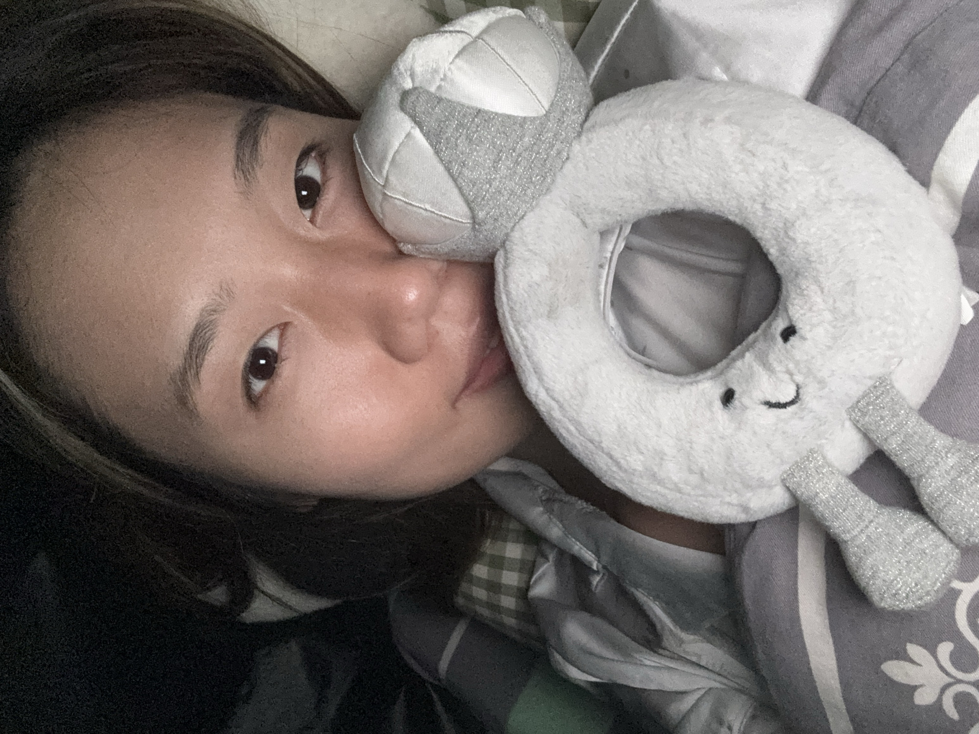 A person with light brown skin and dark brown hair is lying down, turned to their left side, with a white and gray plush toy pressed against their face. The toy appears to be a ring-shaped object with two small protrusions resembling arms or legs and a stitched smile on its front. A smaller, ball-shaped object is attached to the top of the ring. The person's right eye is visible, and their lips are slightly parted. They are lying on a bed with gray and white striped bedding and a gray blanket with a white floral pattern. A green and white checkered pattern is visible on a shirt or blanket underneath the top layer. The location of this image is not discernible from the provided details. Therefore, it is impossible to determine if the location is in Nan'an District, China.