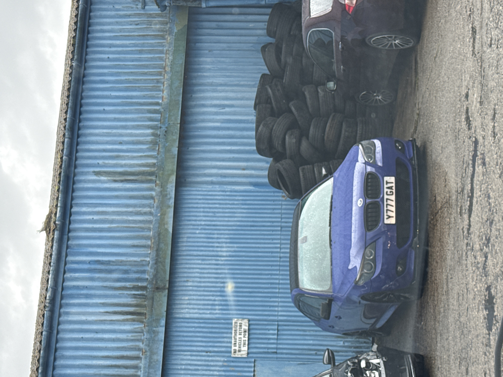 The image depicts an outdoor scene, likely an industrial or automotive area in Great Yarmouth, United Kingdom. A prominent feature is a tall, blue corrugated metal building that spans most of the background, showing signs of weathering and dampness. The ground in front of it is a rough, dark asphalt or concrete surface, also appearing damp.
In the foreground and mid-ground, several vehicles are parked. A distinctive, vibrant blue sedan, possibly a BMW, is positioned diagonally, with its front facing towards the viewer's right. Its UK license plate, "Y777 GAT," is clearly visible. The car's exterior, particularly the windshield and hood, appears wet, suggesting recent rain or condensation. To the right of the blue car, a darker-colored hatchback is partially visible, with its multi-spoke silver wheels prominent.
Between the parked vehicles and the blue building, a large collection of black tires is stacked high, forming a significant mound.
The sky, visible in the upper left, is overcast, indicating a cloudy daytime setting. A small white rectangular sign with black text is affixed to the blue corrugated wall, reading: "NO UNAUTHORISED VEHICLES BEYOND THIS POINT," suggesting a restricted or private area. No people are visible in the scene. The overall impression is of a utilitarian outdoor space used for vehicle or tire storage.