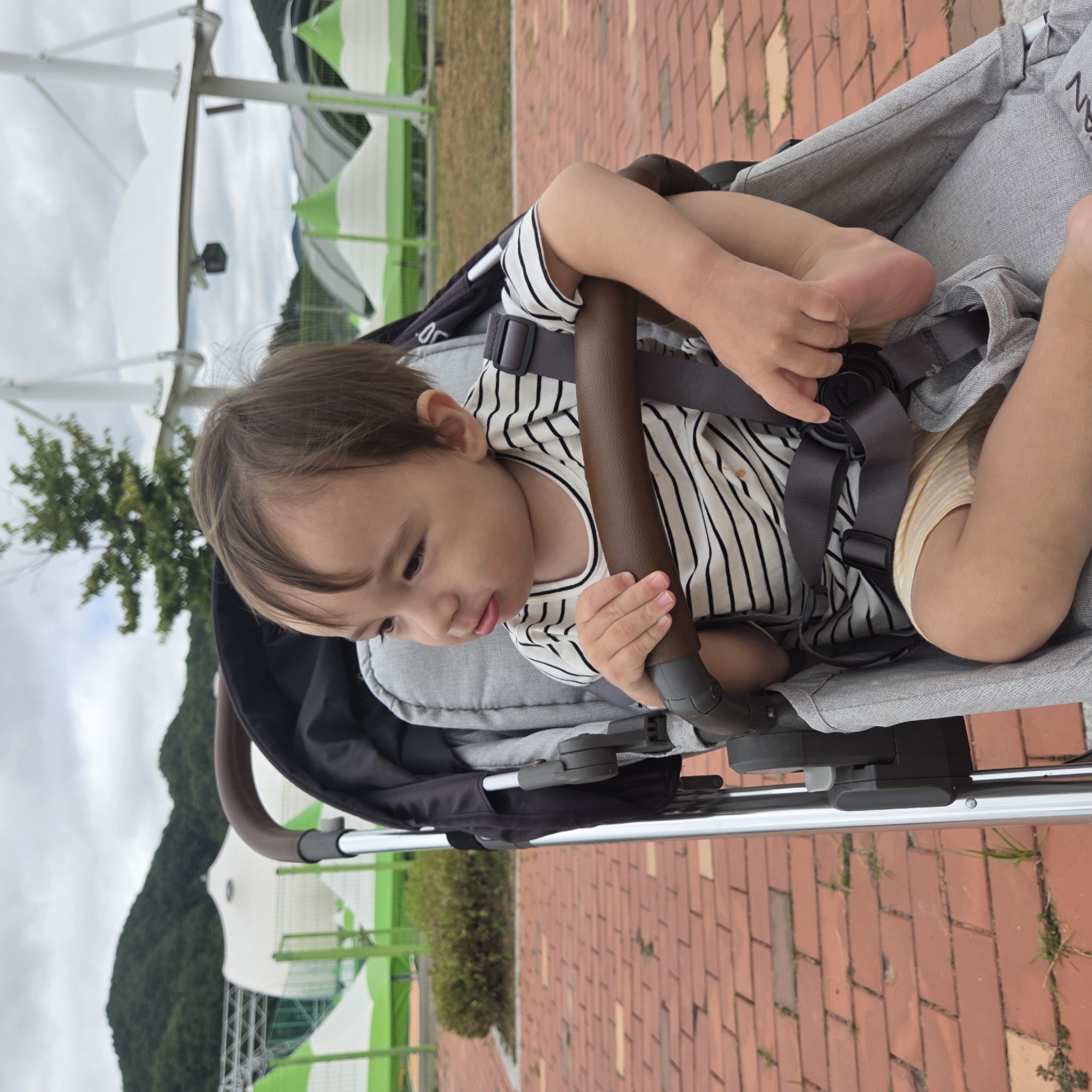 A young child sits in a stroller, facing left, with their right foot propped on the stroller's safety bar. The child is wearing a black and white striped t-shirt and yellow and white striped shorts. The stroller is made of gray fabric and has a brown handlebar. The background shows a paved area with red bricks, green bushes, and several white and green tents or canopies under a cloudy sky. The image appears to have been taken outdoors.