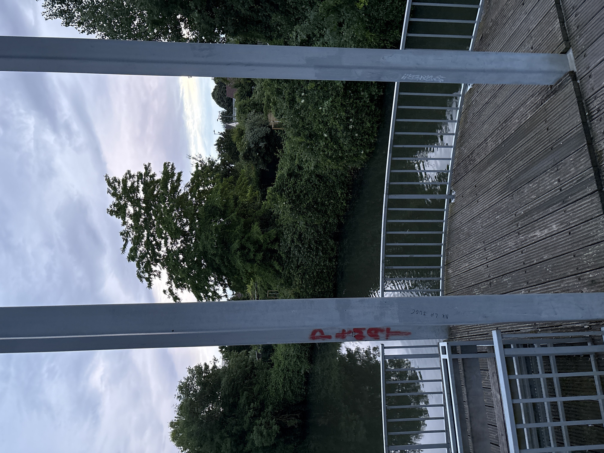 View from a metal bridge looking out at a tree-lined river in Unknown, Unknown. The bridge has a wooden deck and metal railings. Red graffiti is visible on one of the bridge's support beams. The sky is overcast.