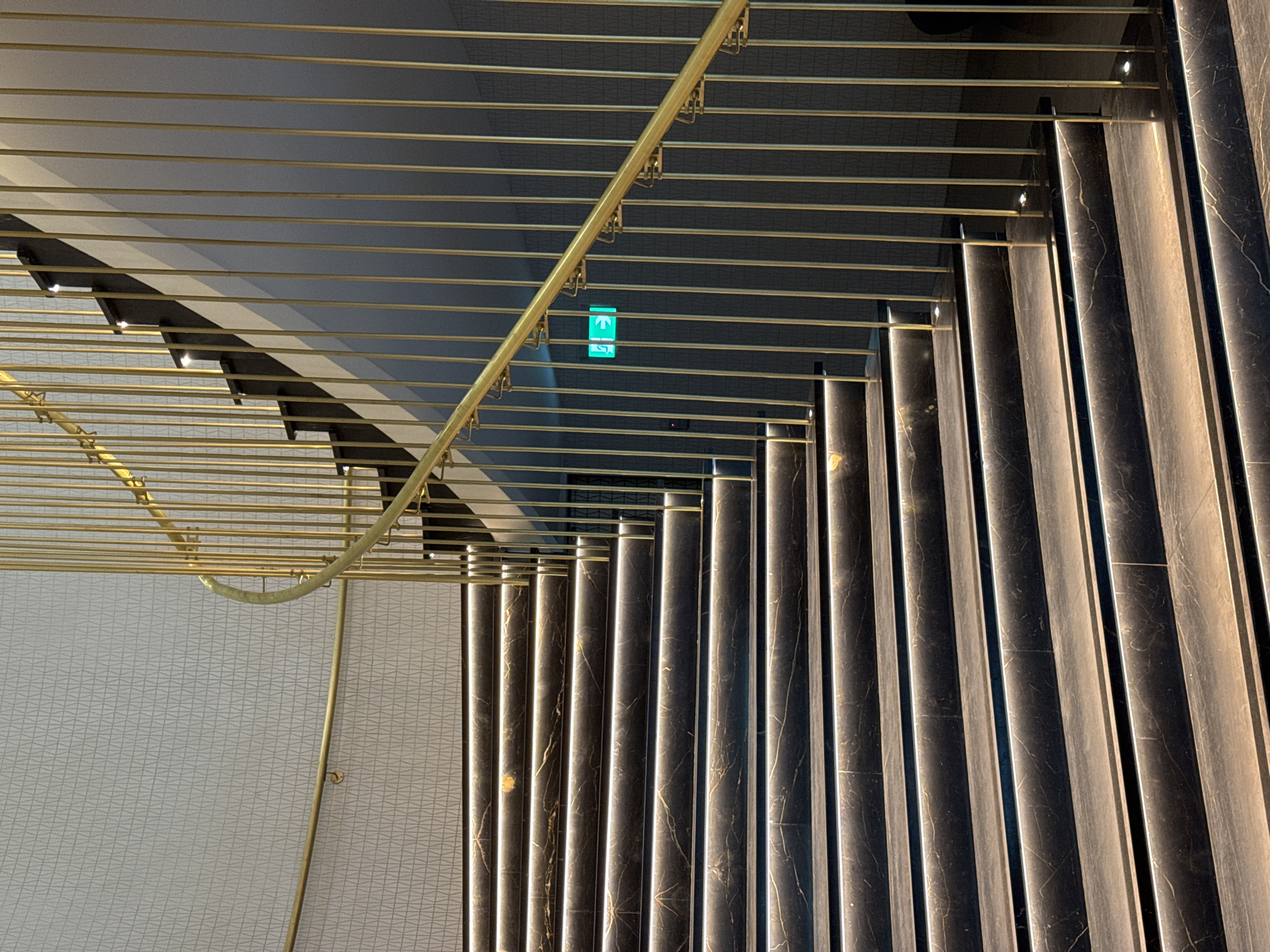 The image depicts an interior staircase in an unknown city. The staircase is constructed with dark, marble-like vertical slabs. The walls are covered with a light geometric pattern. A golden handrail runs along the stairs. An exit sign with a green arrow is visible.