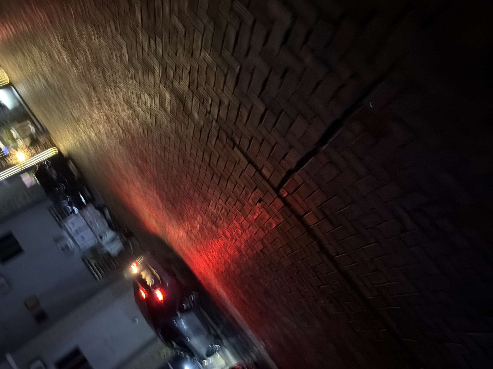 In Bukuru, Nigeria, at night, the red taillights of a dark-colored vehicle are illuminated, casting a vibrant glow on the herringbone-patterned brick pavement. The vehicle is in motion, evidenced by the slight blur. Adjacent to the pavement, a building with windows and architectural features is visible, dimly lit. Further back, another vehicle, possibly a light-colored SUV or van, appears to be parked or stationary. The scene is characterized by low light conditions, with artificial lighting from the vehicles and building contributing to the overall illumination. The ground surface appears wet or reflective, enhancing the glow of the vehicle's taillights. The presence of outdoor lighting fixtures with yellow hues is also noted in the background.