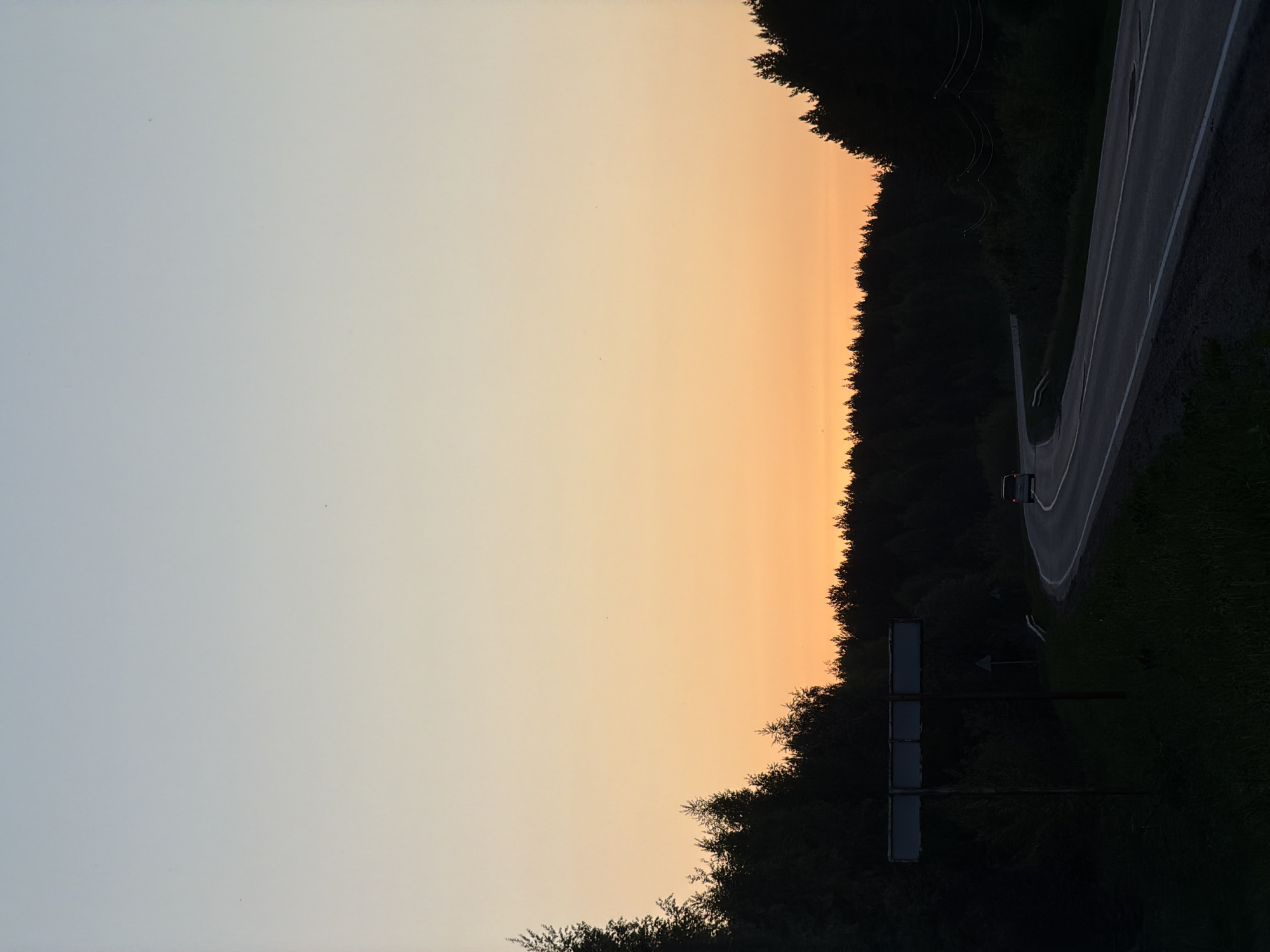 A landscape oriented photo depicts a rural setting near Oreshniki, Russia. The sky transitions from light blue to orange near the horizon. A dense forest line divides the sky from a roadway that curves to the right. A sign structure is present at the bottom left of the forest area. On the road, a single vehicle with illuminated tail lights is driving along the road.